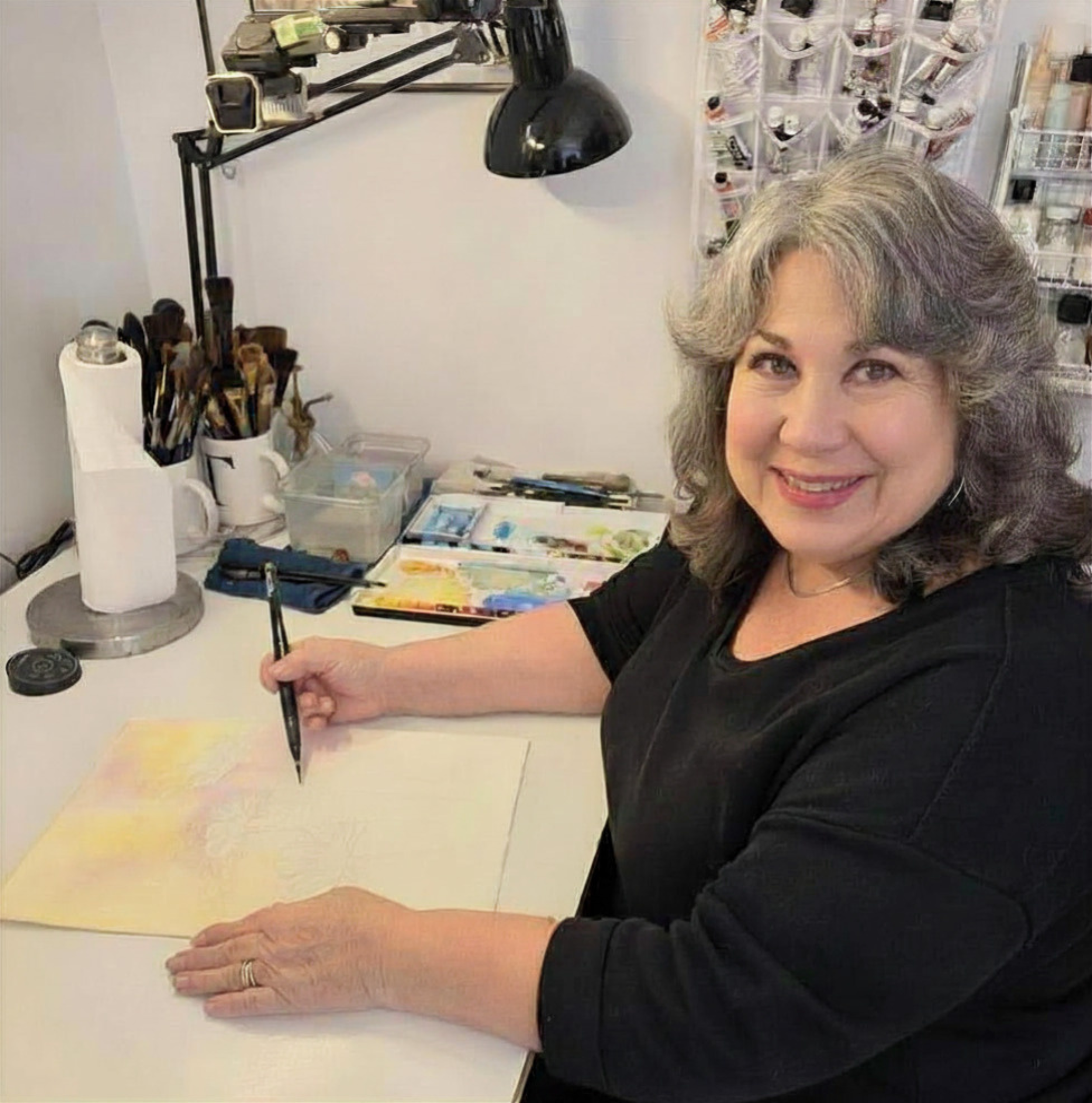 Lorraine Rimmelin in her watercolor art studio. The workspace includes art supplies, paints, brushes, and a watercolor painting on the table. In the background, there are organized art supplies and shelves.