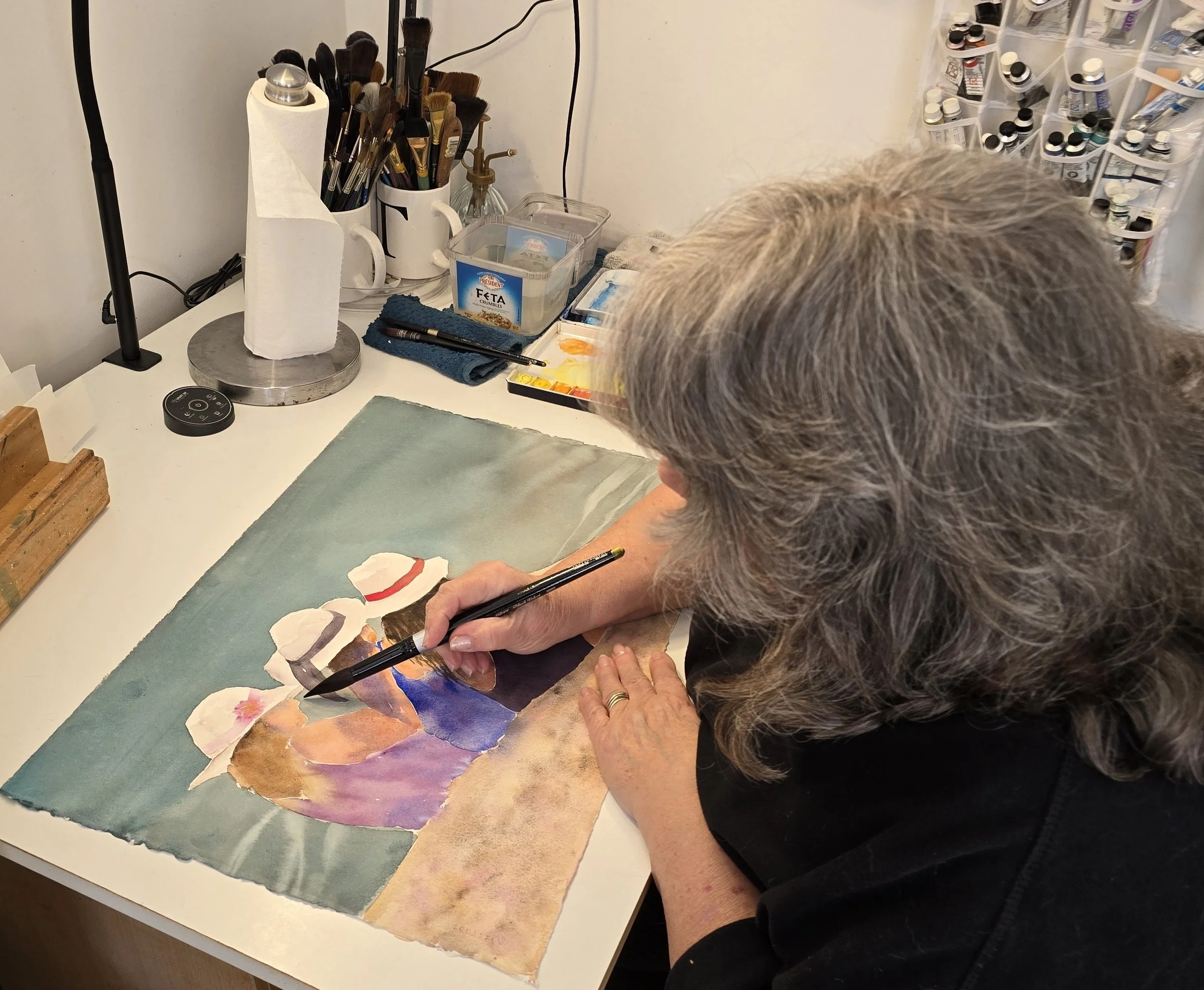 Lorraine Rimmelin working on a watercolor painting at a white table with art supplies around.