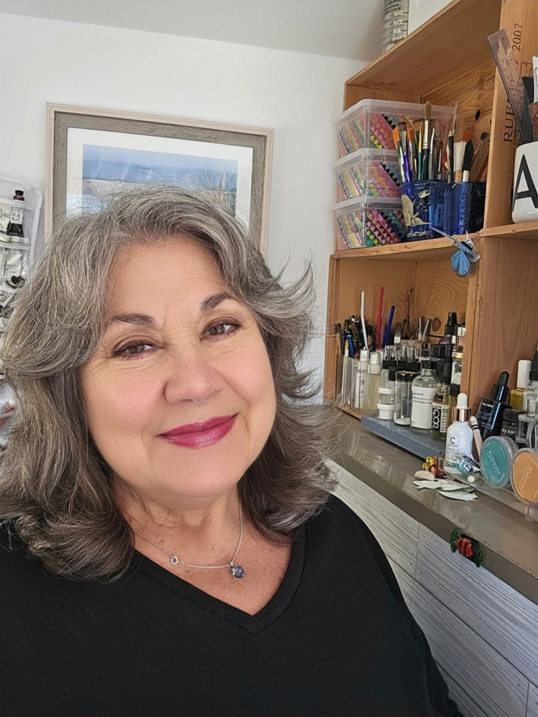 Lorraine Rimmelin, watercolor artist and teacher, with shoulder-length gray hair and pink lipstick smiling in a room with a wooden shelf filled with art supplies and a framed picture on the wall.