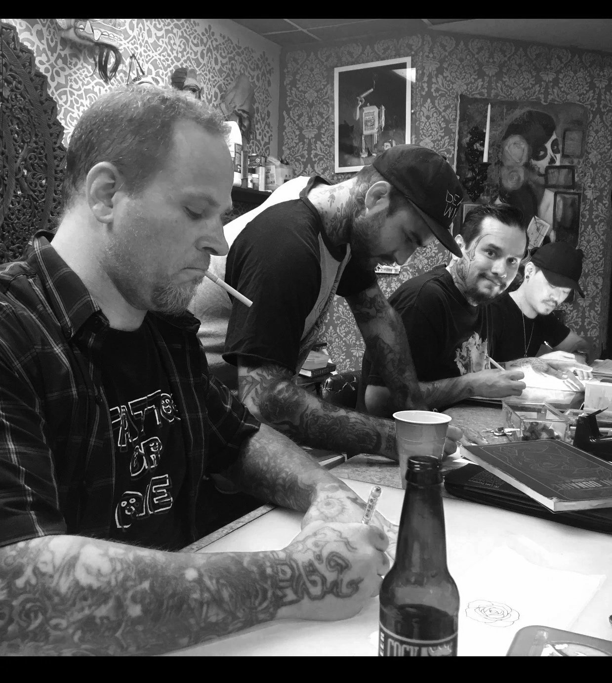 Four men with tattoos sitting at a table, sketching or drawing, in a room with patterned wallpaper and artwork on the wall.