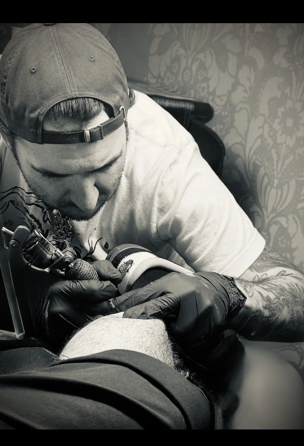 A man with tattoos on his arms, wearing a baseball cap and black gloves, getting a tattoo on his leg from a tattoo artist.
