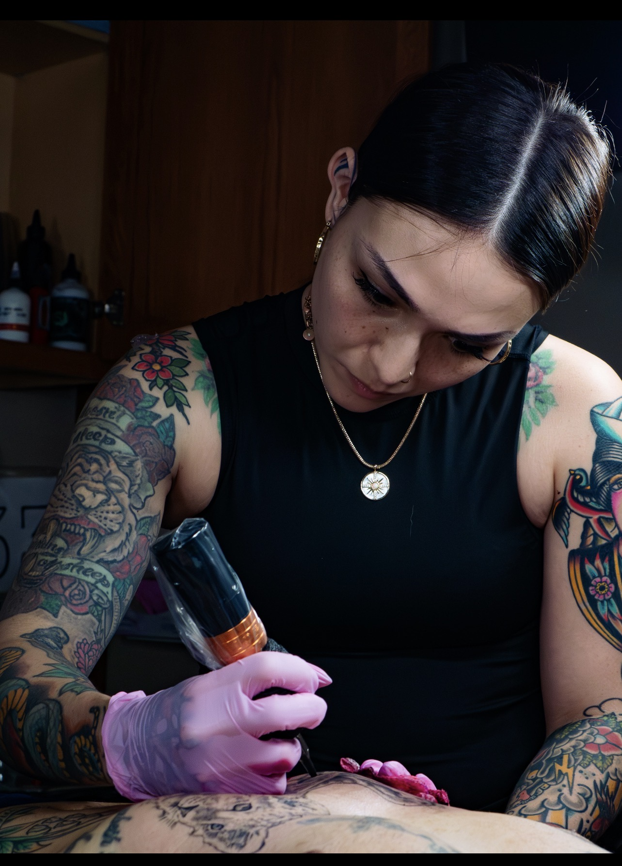 A woman with dark hair, tattoos on her arms, and wearing a black sleeveless top is tattooing a person's arm. She is wearing pink gloves, holding a tattoo machine, and looking down at her work.