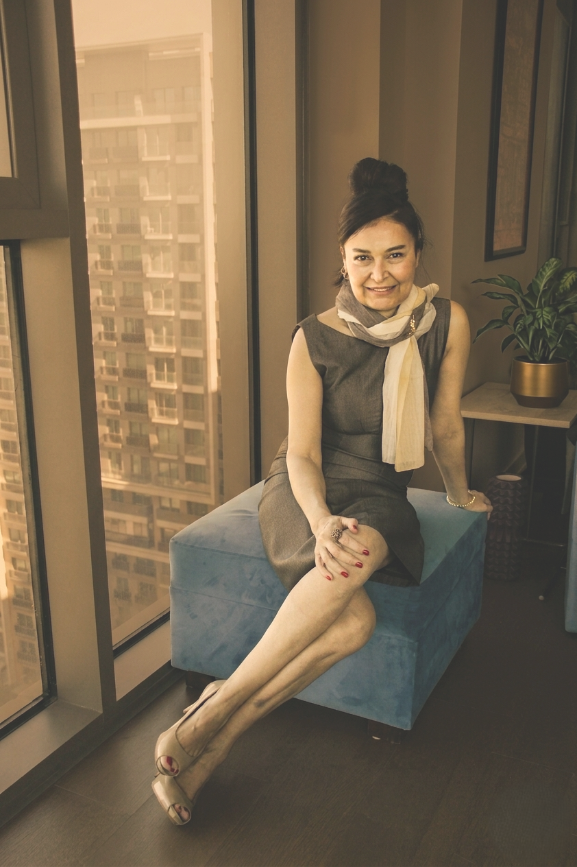 A woman sitting on a blue ottoman near a large window in a modern apartment, smiling at the camera. She is wearing a sleeveless gray dress, sheer stockings, beige high heels, and a scarf around her neck. A green plant is on a side table behind her.