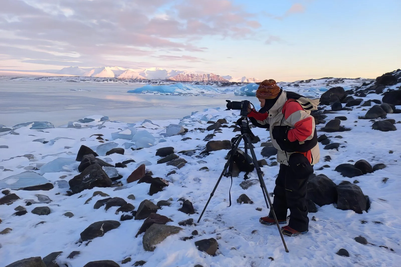 Person in winter clothing setting up a camera on a tripod on a snowy, rocky shoreline near a frozen body of water with snow-covered mountains in the background.