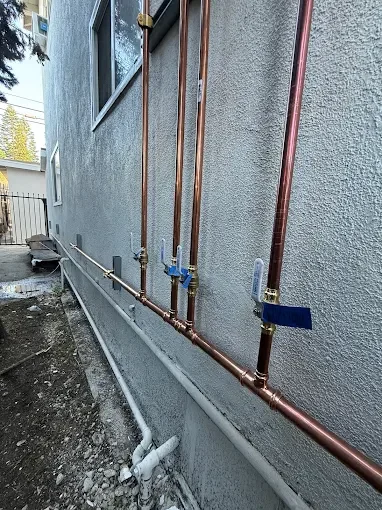 Copper plumbing pipes mounted on an exterior wall with some valves and fittings, near a house foundation.