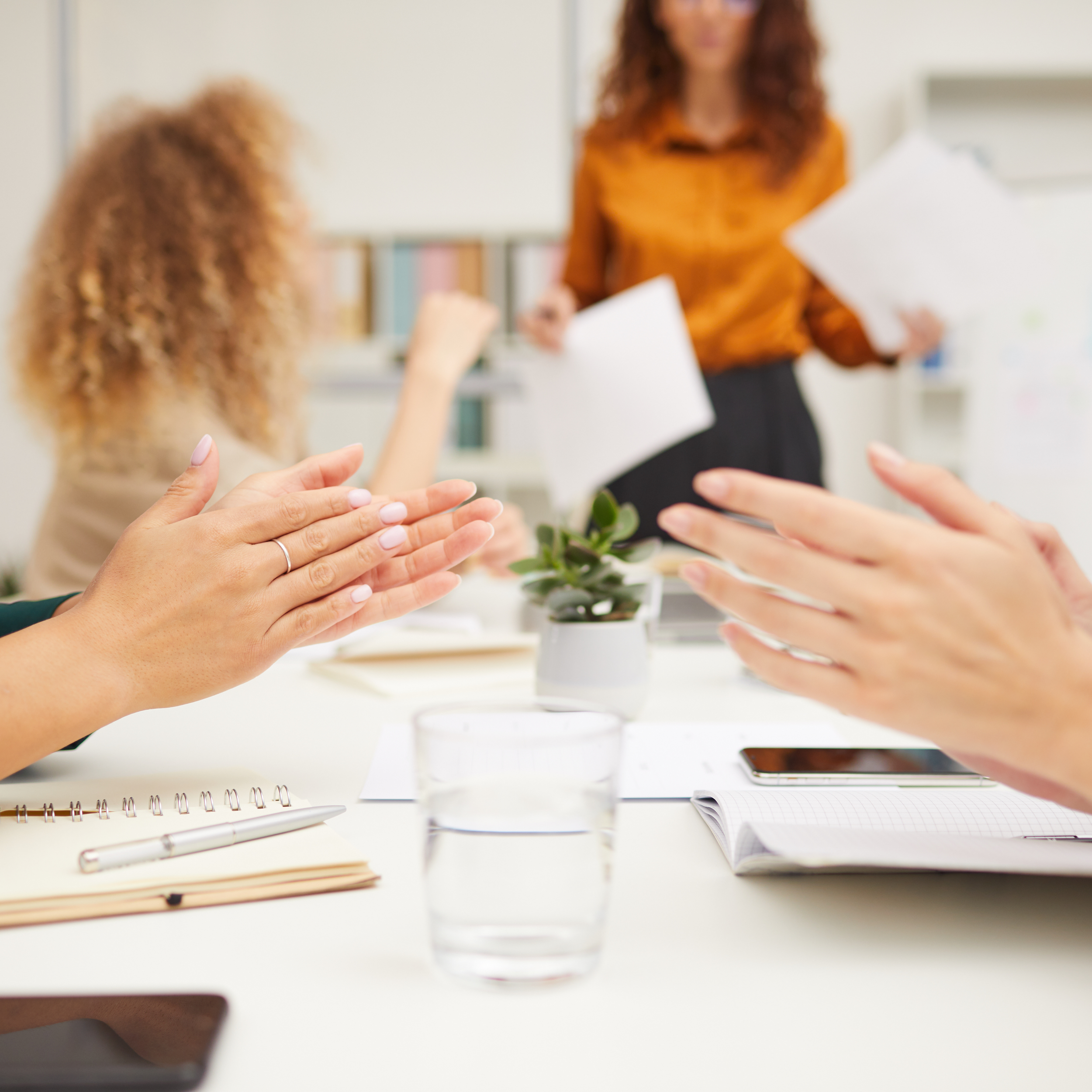A group of coworkers sitting around a table clap during a meeting while a woman stands in the background holding papers, appearing to present or lead the discussion. A notebook, smartphone, glass of water, and a small potted plant are on the table.