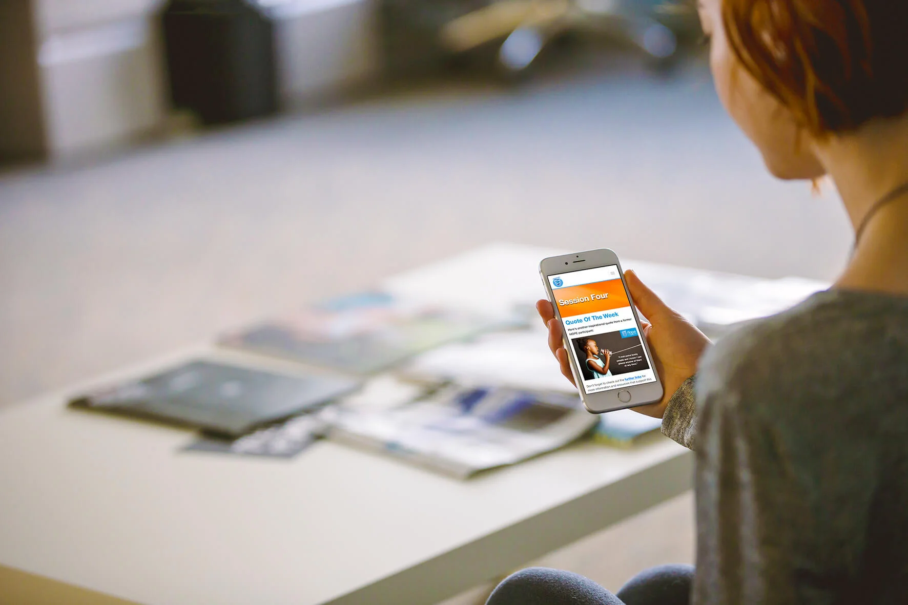 A woman with short hair holding a smartphone displaying a presentation slide titled "Session Four" with a subtitle "Quote Of The Week". She is sitting at a table with various pamphlets or flyers spread out on it. The background is blurred.