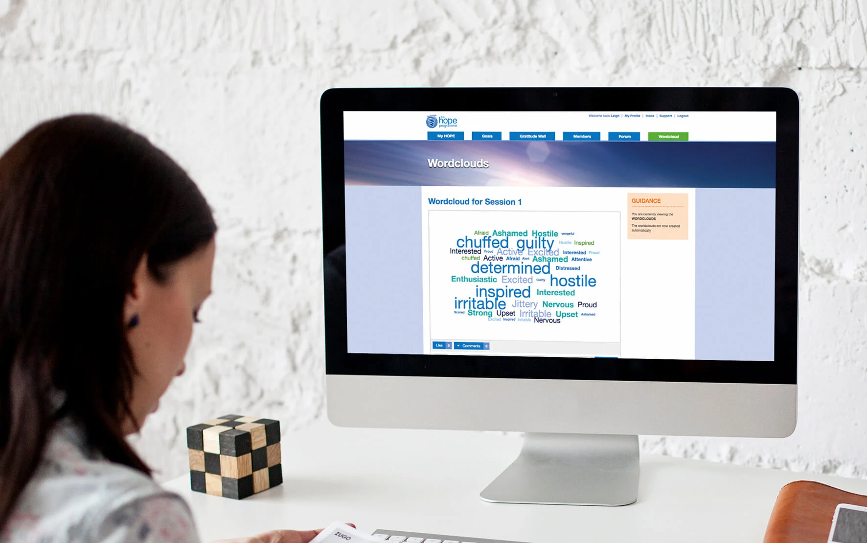 A woman sitting at a desk using an iMac computer. The screen displays a word cloud with words like "guilty," "hostile," "inspired," and "irritable." A black-and-wooden cube puzzle is on the desk, with white textured walls in the background.