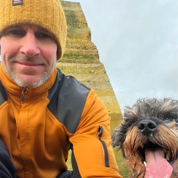 A man wearing a yellow beanie and an orange jacket taking a selfie outdoors with a happy, panting dog with a scruffy gray and brown coat.