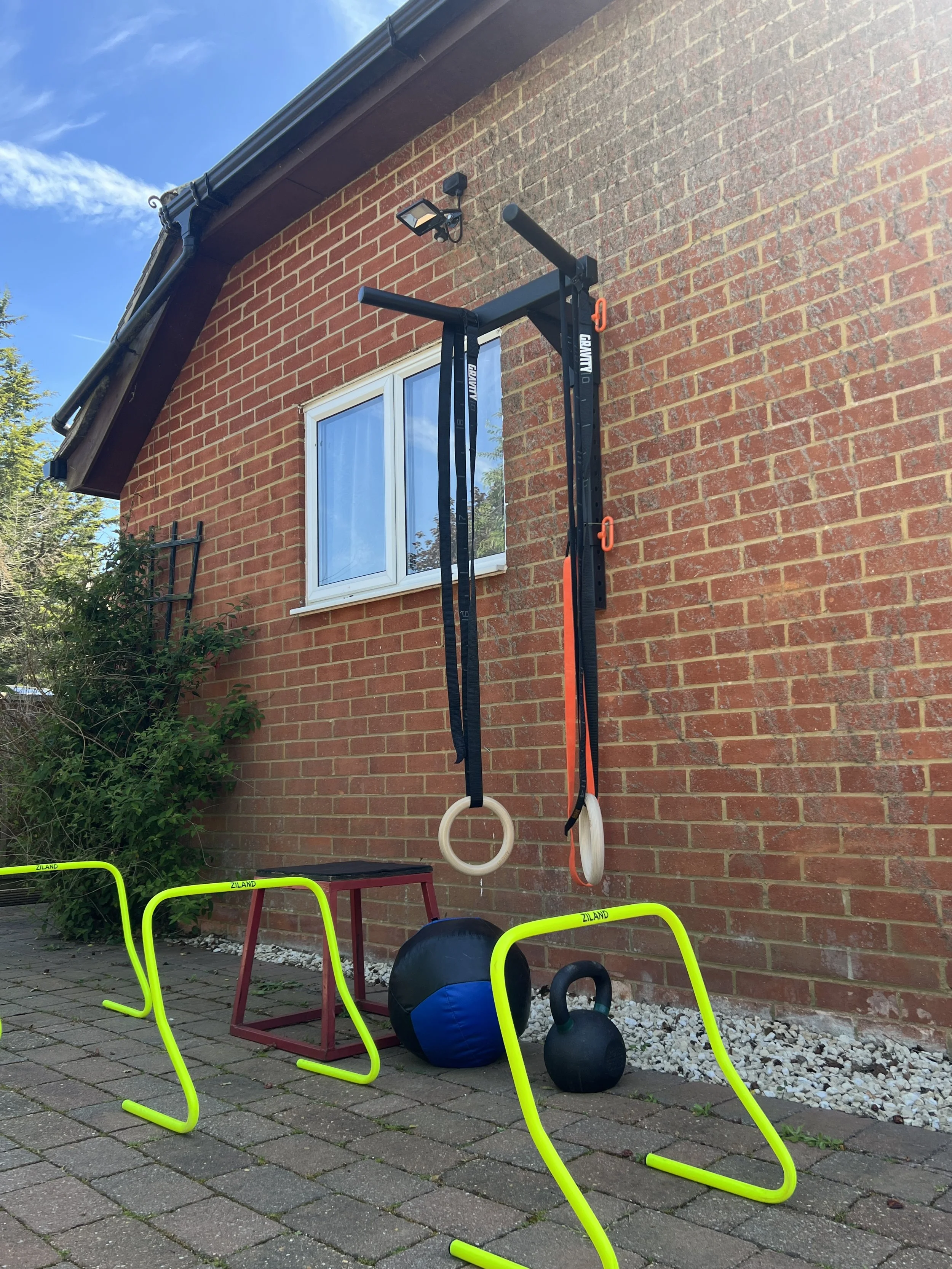 Outdoor fitness area with gymnastic rings, kettlebells, agility hurdles, and a medicine ball set up on a brick patio beside a red brick house with a window, greenery, and a blue sky.