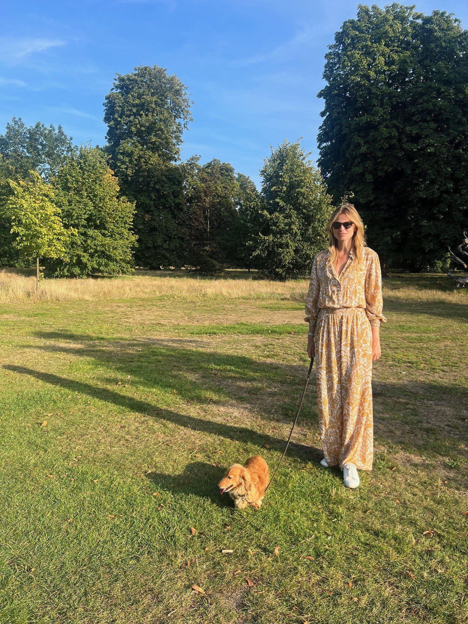 A woman with blonde hair wearing sunglasses, a patterned long dress, and white sneakers standing on grass in a park with trees in the background. She is holding a leash attached to a small, fluffy, golden-colored dog with its tongue out.