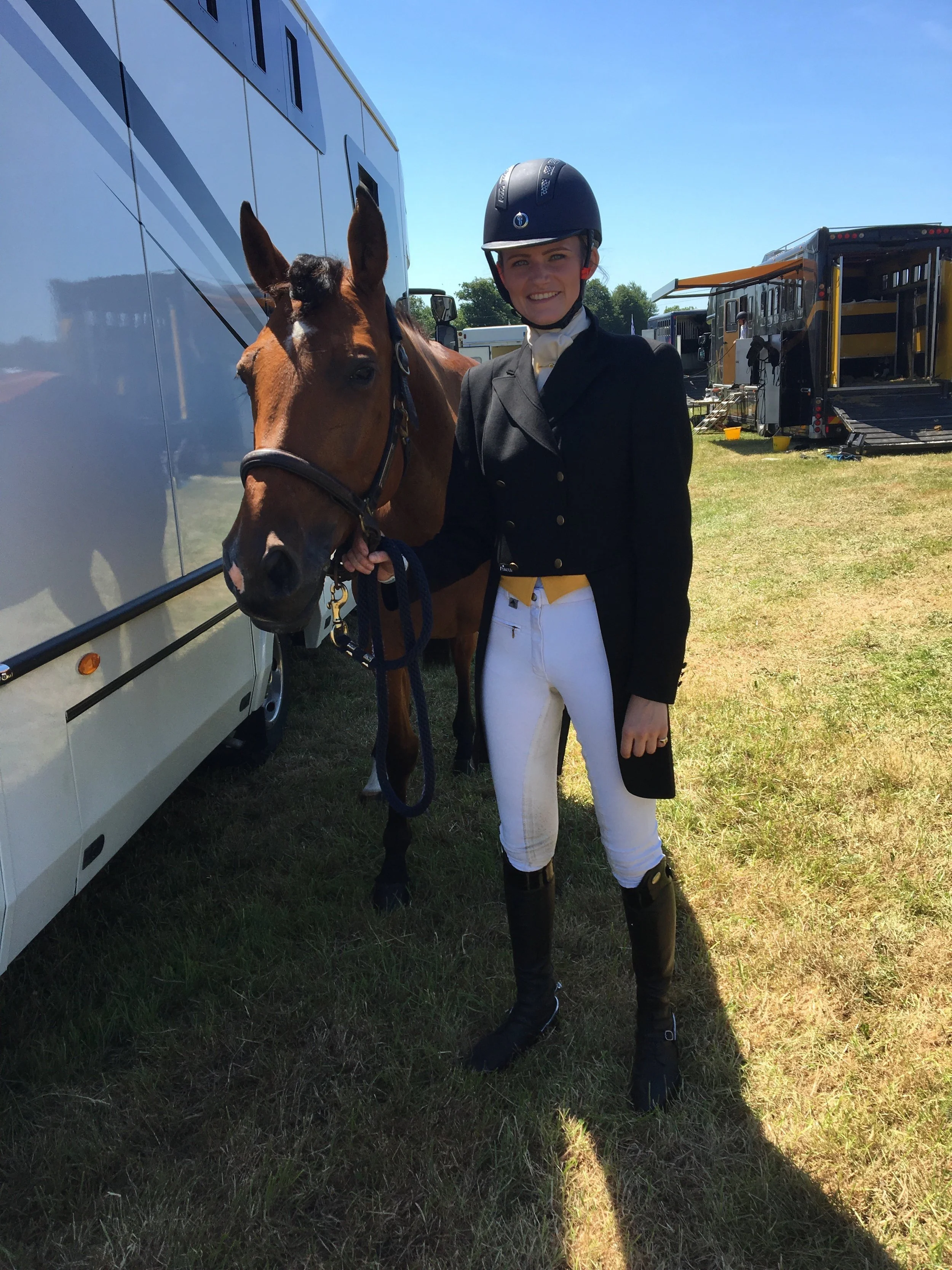 A smiling female equestrian dressed in riding black jacket, white breeches, and black riding boots, wearing a black helmet, standing outdoors on grass with a brown horse. A large horse trailer and other equipment are in the background on a sunny day.
