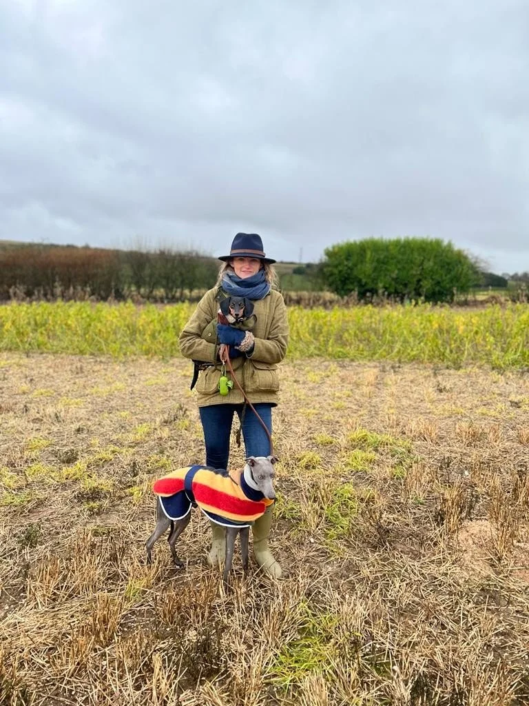A woman standing in an open field holding a small dog, wearing a hat, scarf, and jacket, with another dog on a leash wearing a colorful sweater.