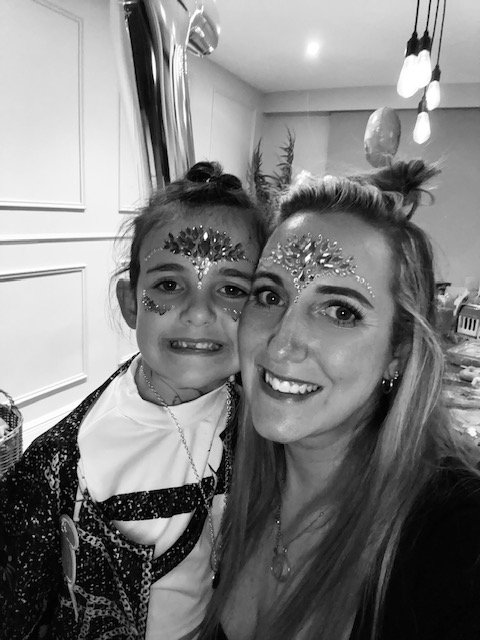 A woman and a girl smiling with face decorations and glitter, taking a selfie together indoors.