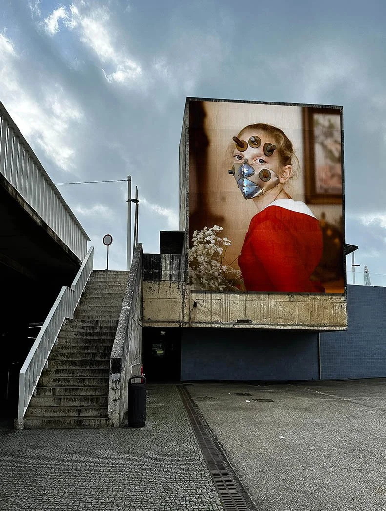 Large billboard on a building showing a woman in a red dress with a face mask and several screws or bolts on her face. The billboard is located outdoors near a set of concrete stairs and a parking lot.