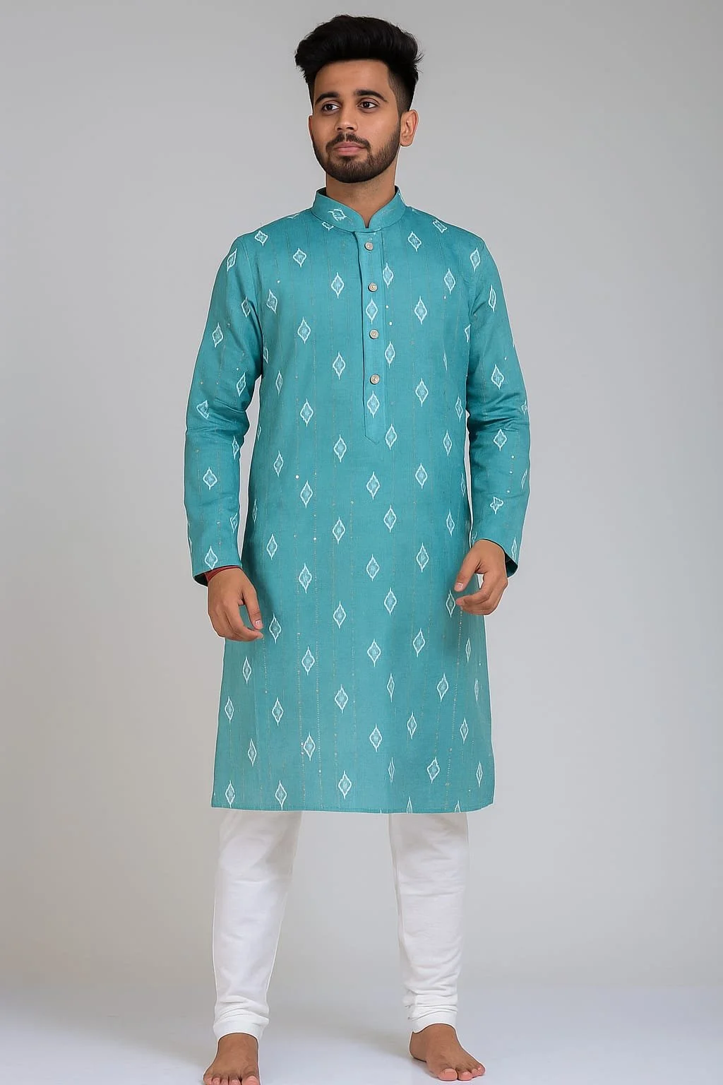 Young man wearing a traditional teal kurta with white patterns and white churidar, standing barefoot against a plain background.