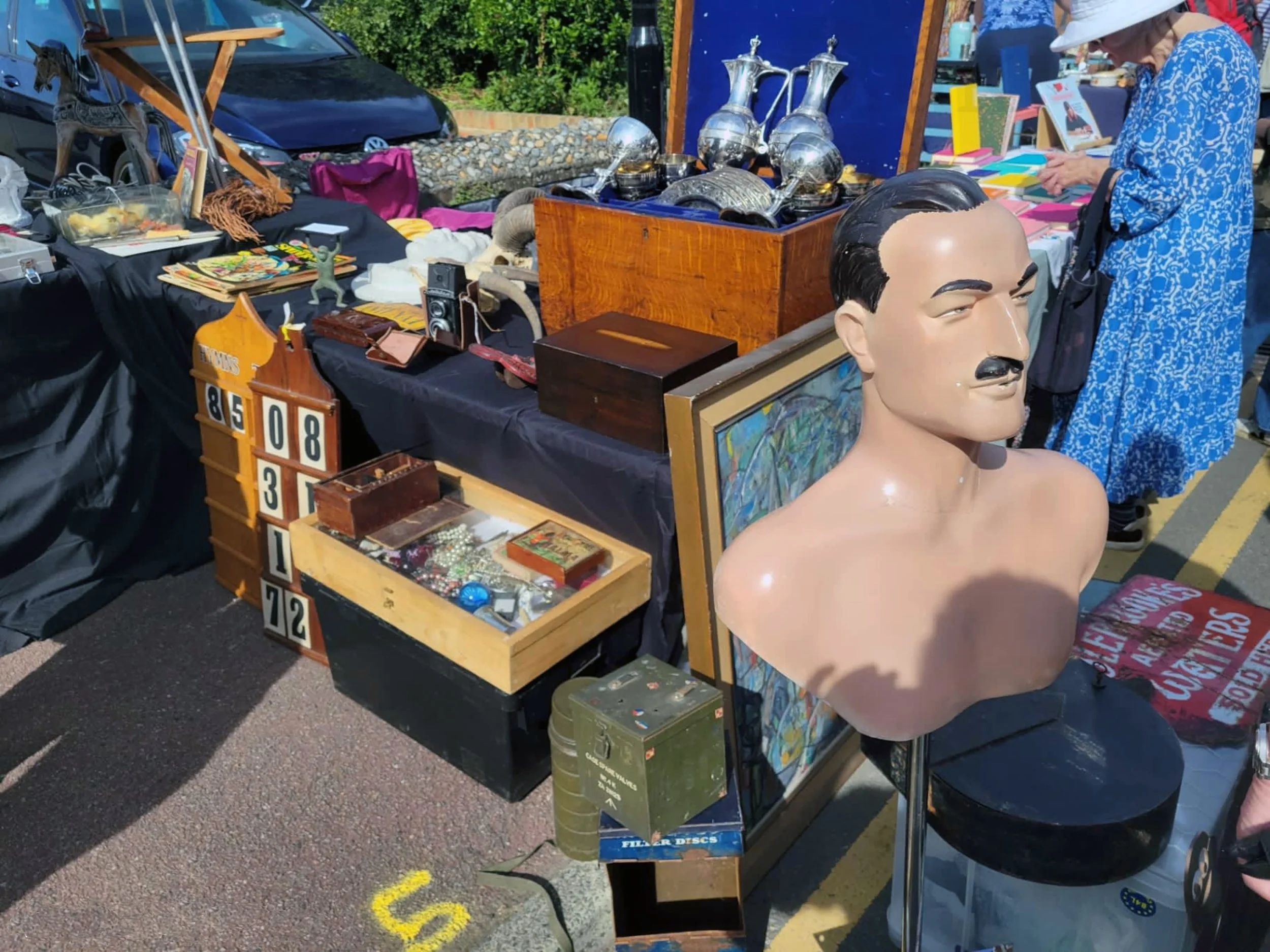 A vintage flea market with tables displaying various collectibles and decorative items, including silver pitchers, a mannequin head, toy soldiers, and books. A woman in a blue dress and wide-brim hat browses the items.