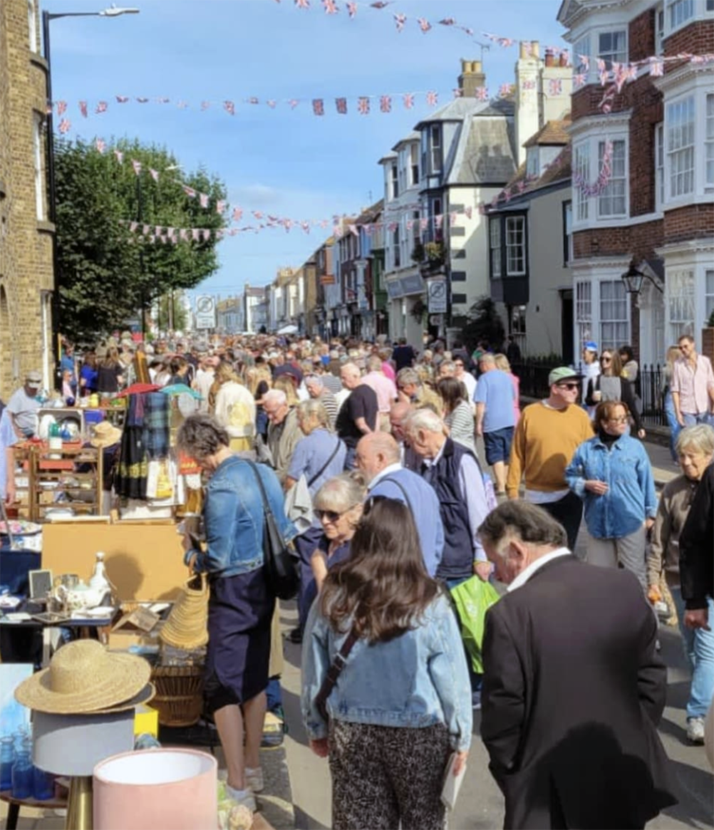 A crowded outdoor street market with vendors and shoppers visible, and bunting hanging overhead.