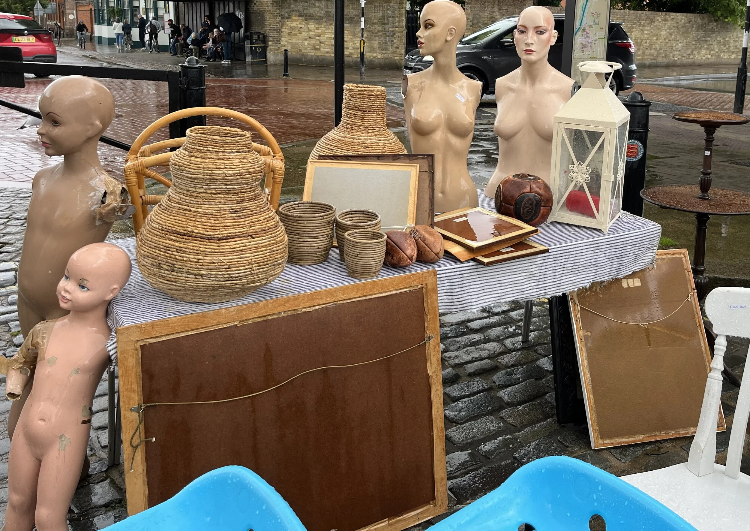A street market stall displaying mannequin busts, woven baskets, framed pictures, an old boxing glove, and a lantern on a table with a striped tablecloth. The scene is wet from recent rain, with people sitting and walking on the cobblestone street in the background.