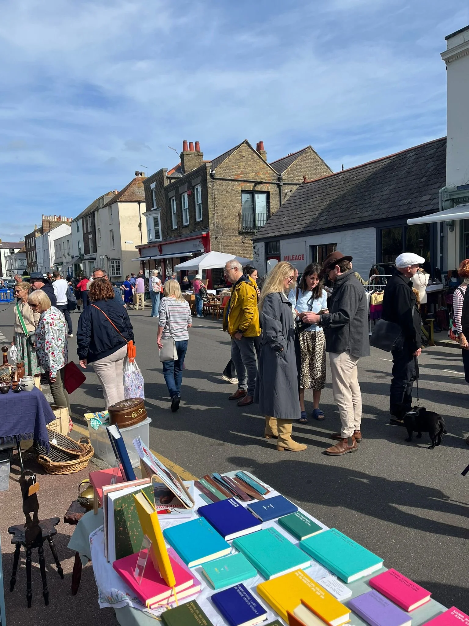 People browsing street market with tables of books, vendors, and shops on a busy street with buildings and blue sky in the background.