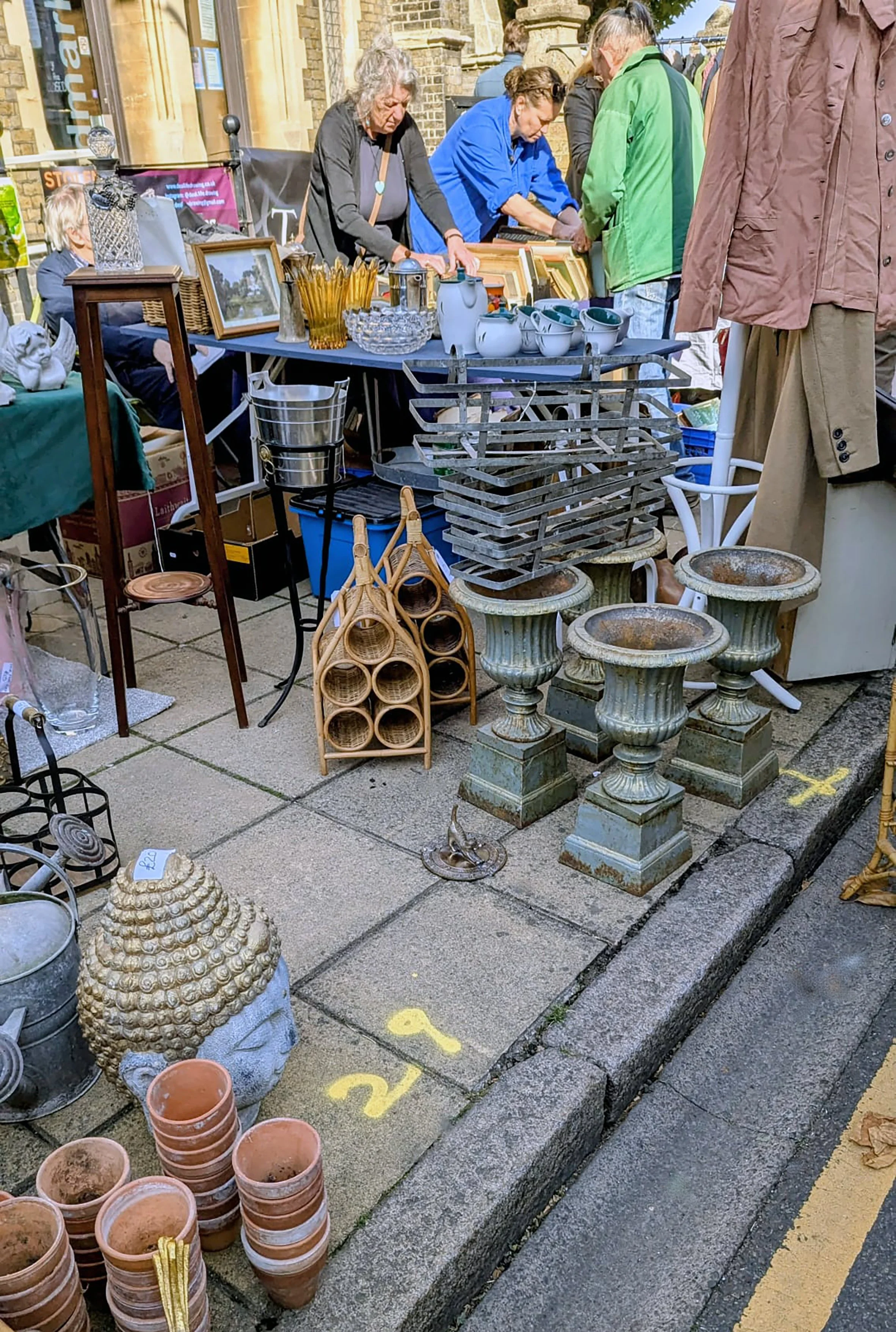 A street market stall with various items, including pottery, decorative sculptures, picture frames, and miscellaneous vintage objects, set up on a sidewalk with people browsing.