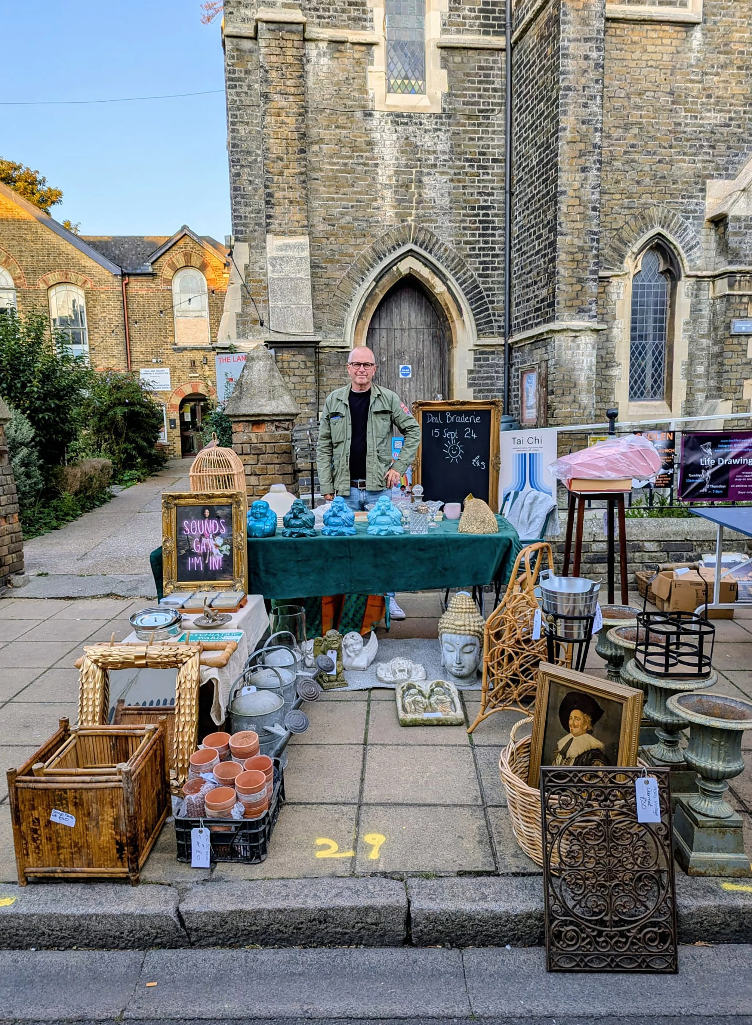 A man stands behind a table of various decorative items such as statues, pottery, and picture frames, set up outside a church with brick walls and stained glass windows. There are other tables with items on the sidewalk, and signs indicating sale or event details.
