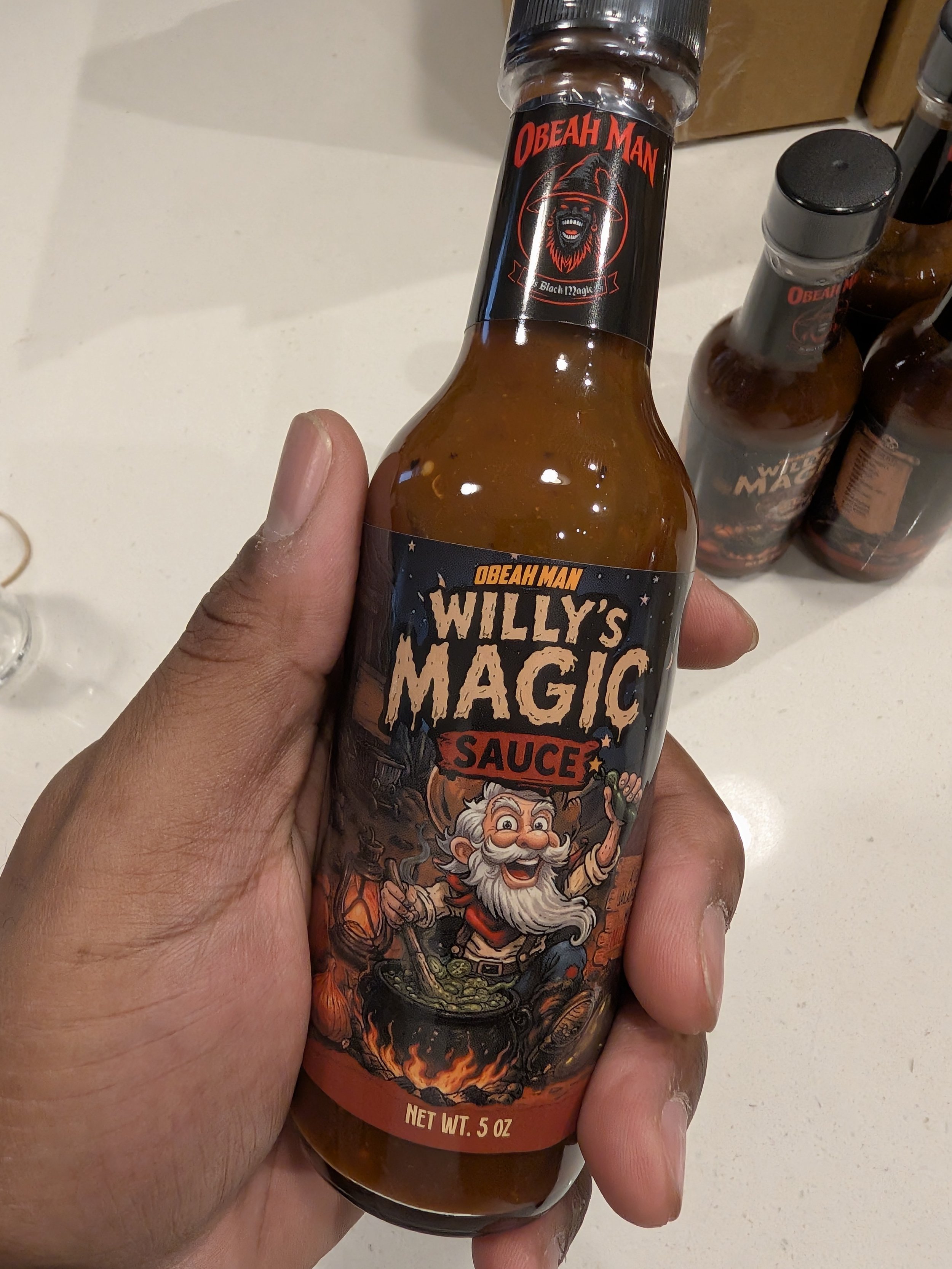 A hand holding a bottle of Willie’s Magic Sauce with a label featuring a prospector with a beard, a hat, and a pot with fire underneath, and with additional bottles in the background.