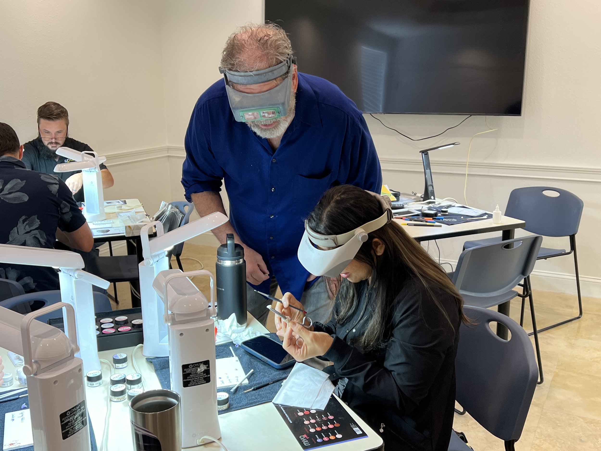 A man with gray hair and a beard wearing magnifying glasses and a blue shirt working with a young woman who has long dark hair and is wearing a black jacket with glasses. They are seated at a table with various medical or laboratory supplies, including vials, a tablet device, and tools. Other people are working in the background, and a large screen is mounted on the wall.