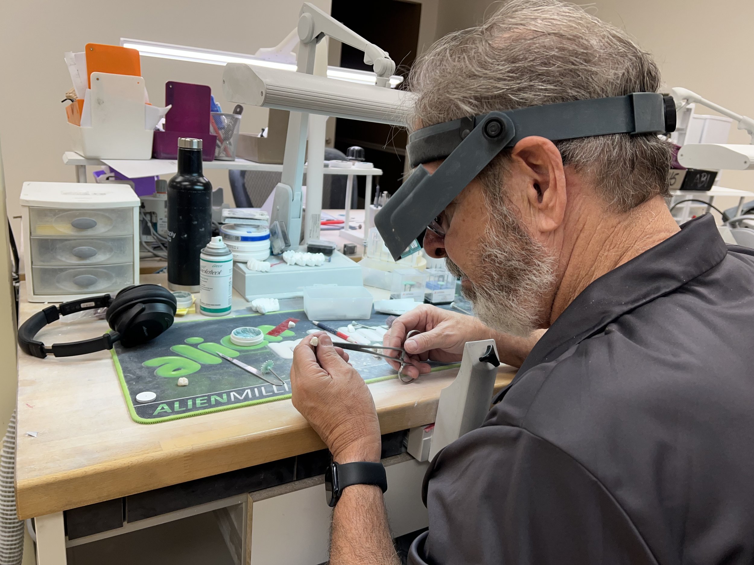 A man with gray hair and a beard wearing magnifying glasses, working at a cluttered workbench with modeling supplies and tools in a workshop or lab environment.