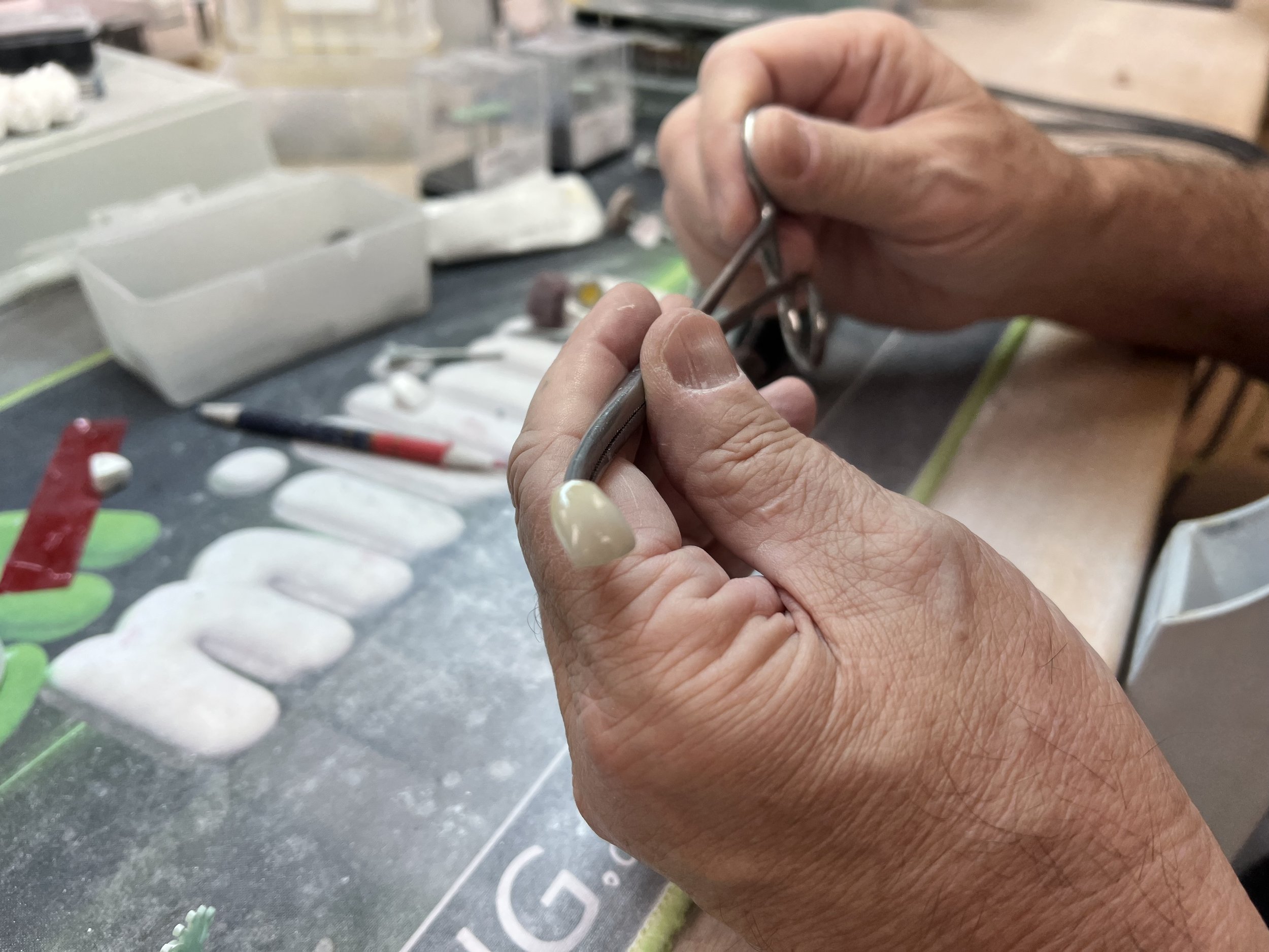 Close-up of a person's hands working with a small metal tool, possibly crafting or repairing jewelry or small metal objects, on a cluttered workbench with various small objects, including white and green items, and plastic containers in the background.