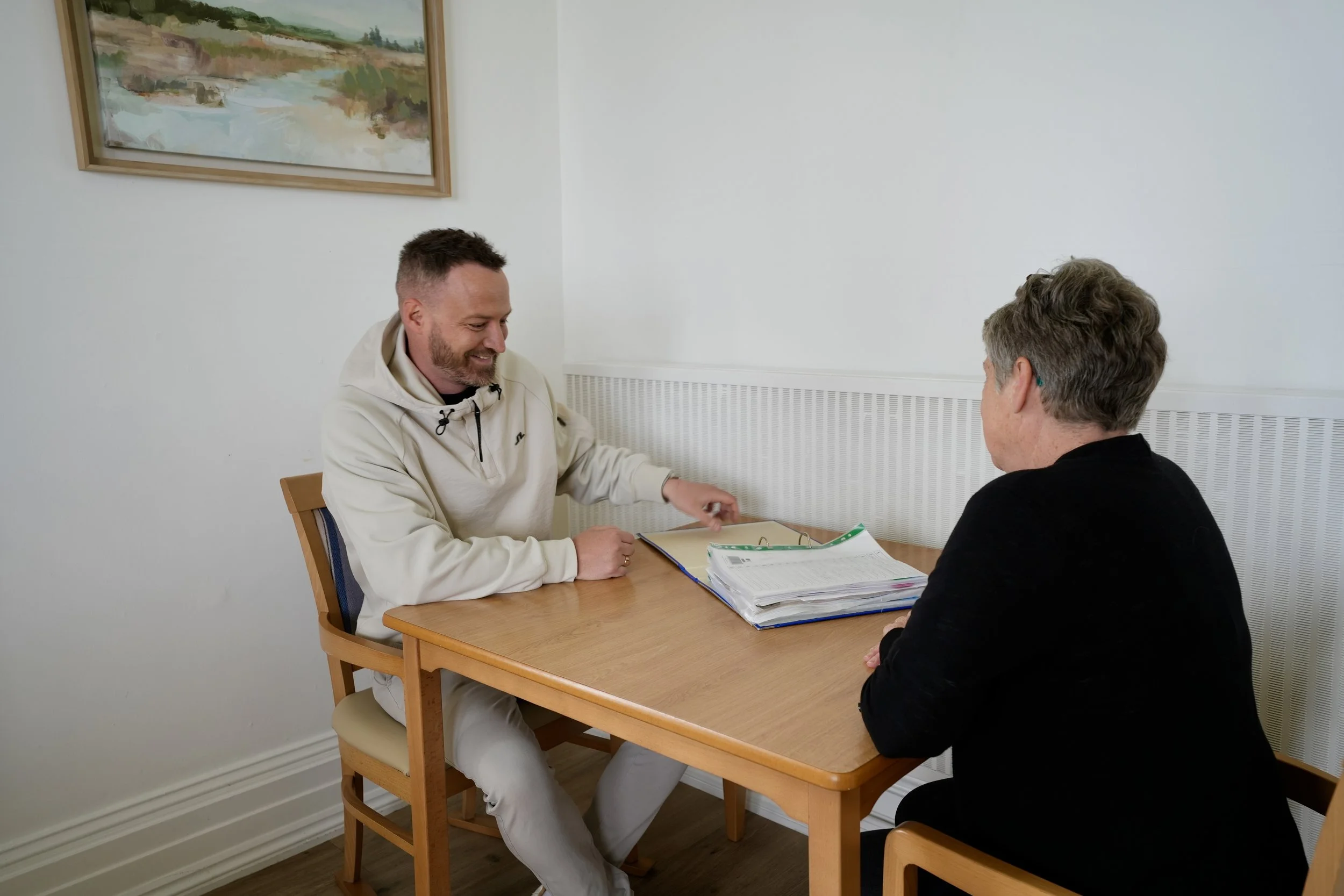 A man and a woman sitting at a small wooden table having a discussion. The man is smiling and pointing at a folder filled with papers, while the woman is listening. There is a framed landscape painting on the wall behind them.