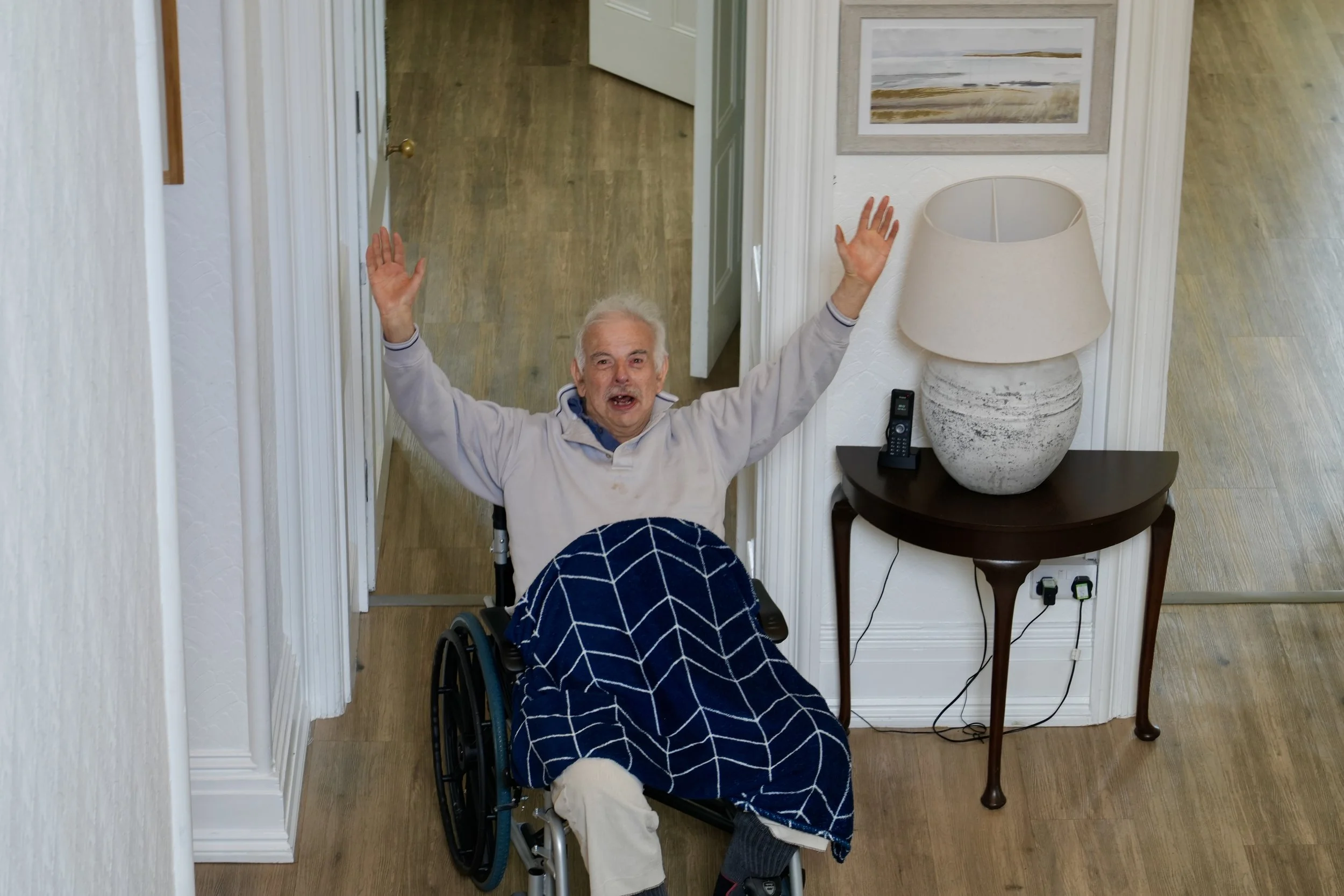 An elderly man with white hair in a wheelchair, raised arms, and open mouth, looking up at the camera in a home interior with wooden flooring, a table with a large lamp, and artwork on the wall.