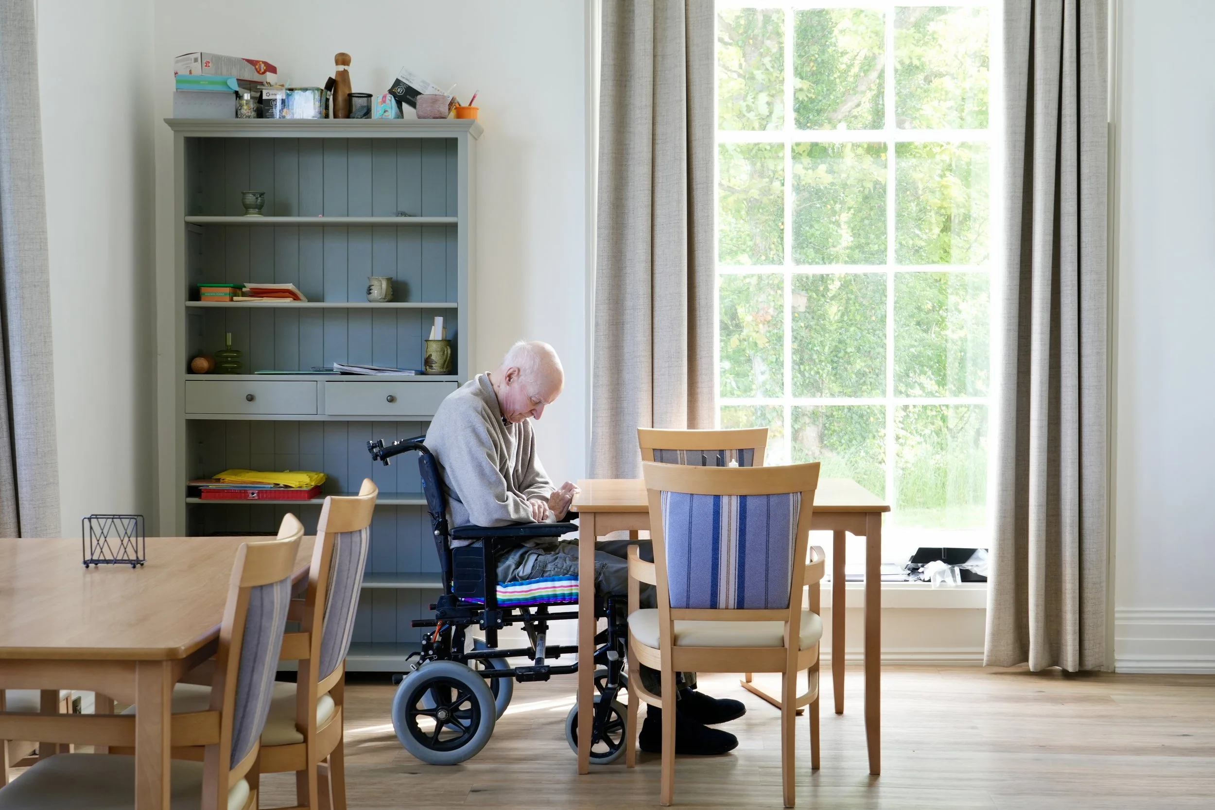 An elderly man in a wheelchair sits at a dining table in a bright room with large windows and curtains, overlooking a green outdoor area.