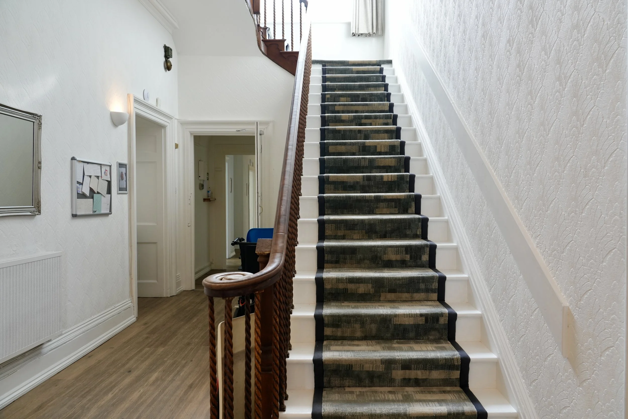 Interior view of a staircase with a wooden handrail, a beige wall with textured wallpaper, a door at the bottom, and natural light coming from a window at the top.
