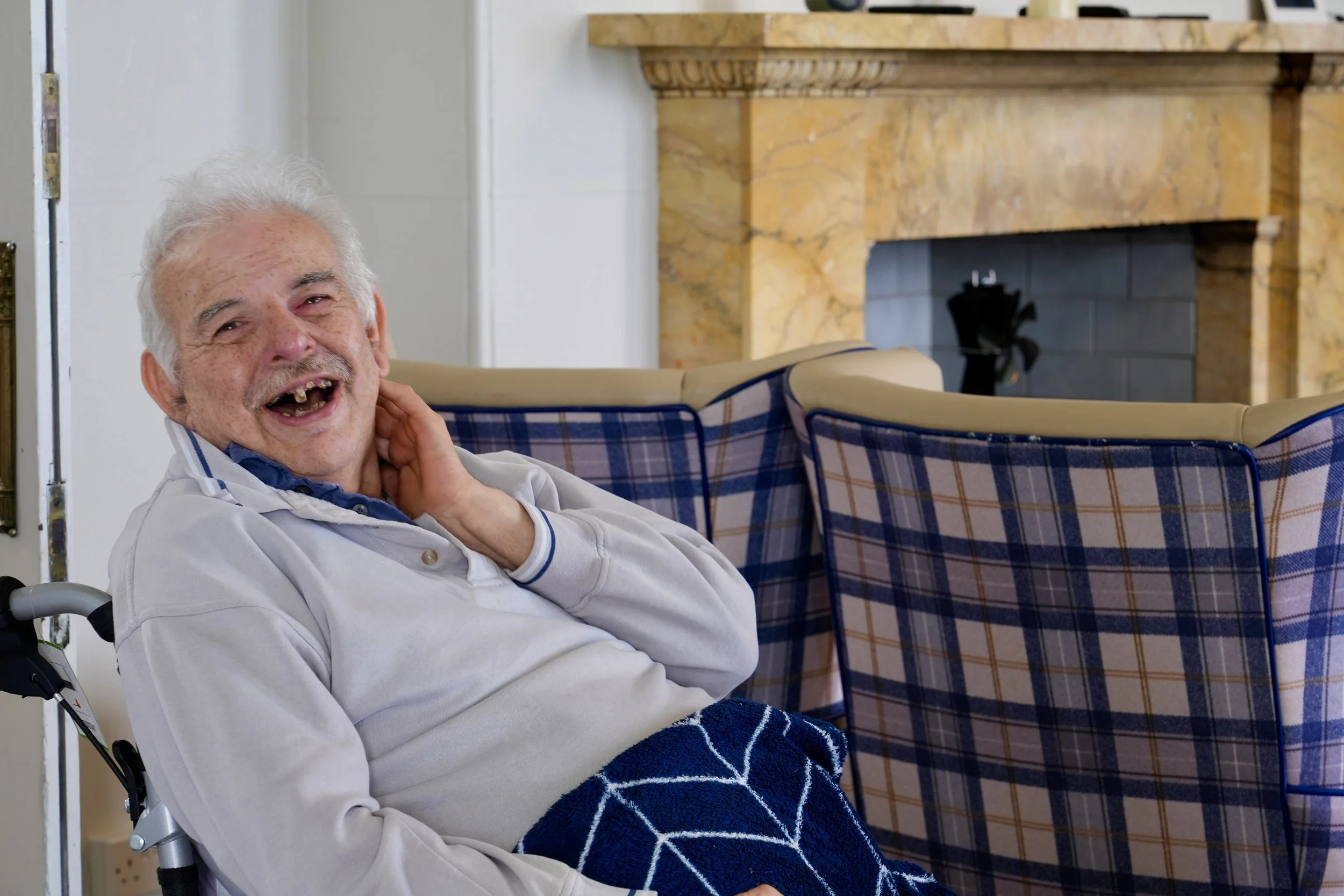 An elderly man with white hair and a big smile sitting in a wheelchair, touching his neck, with a blue patterned blanket over his lap, in a room with a fireplace and plaid upholstered chairs in the background.