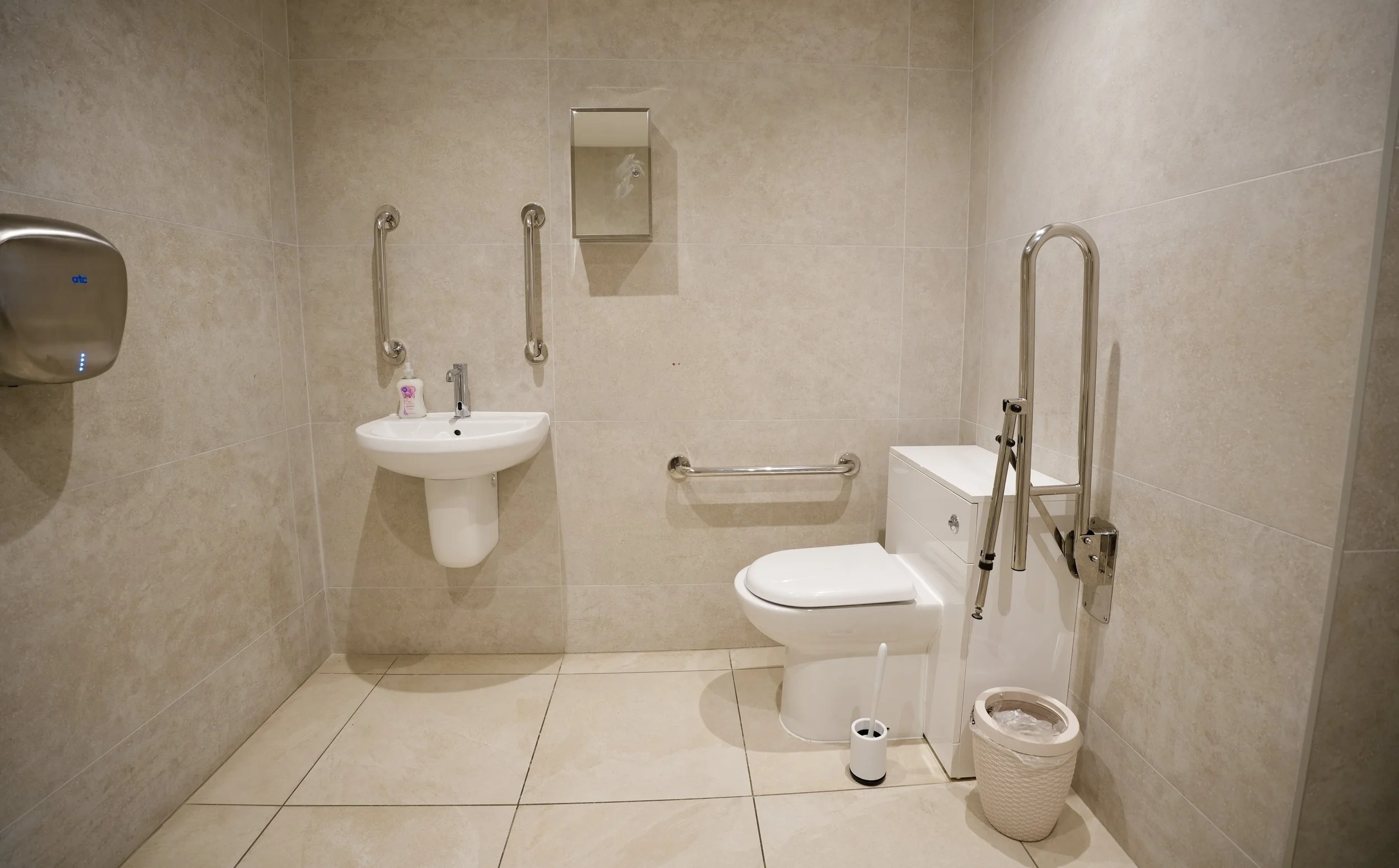 An accessible bathroom with beige tiled walls and floor, featuring grab bars near the sink and toilet, a sink with a soap dispenser, a mirror, a toilet with a raised seat, a ladder, a wastebasket, and a portable air purifier.