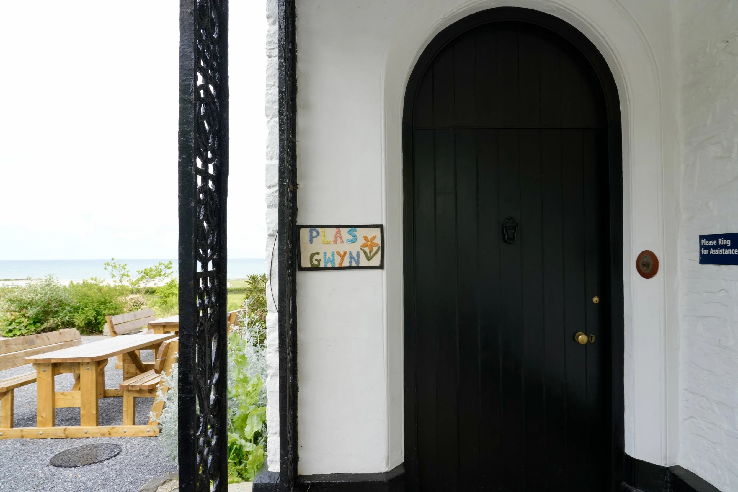 Door with a sign that says 'PLAS GYN' and a flower drawing, set in a white wall, with outdoor seating and greenery visible outside.