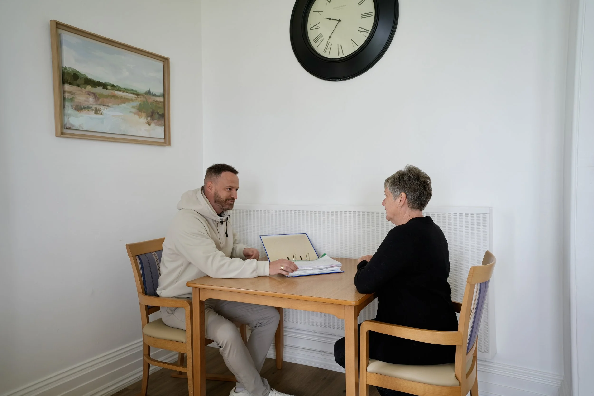 A man and a woman sit at a wooden table facing each other in a plain room with white walls. The man is wearing a white hoodie, and the woman has short gray hair and is dressed in a black top. They are engaged in conversation with a folder or binder o