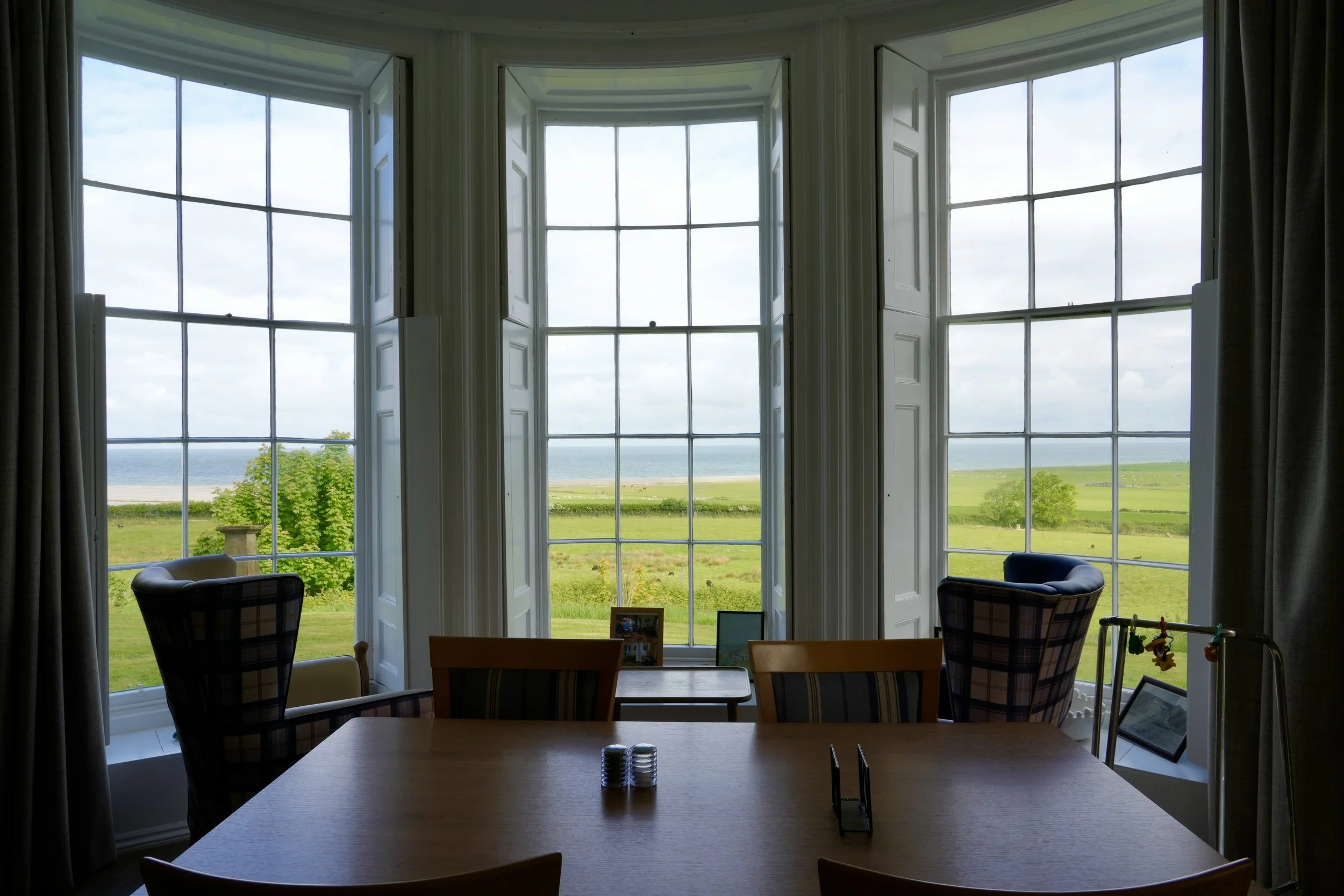 View of a large window overlooking a green landscape with the sea in the distance, taken from inside a room with chairs and a wooden table.