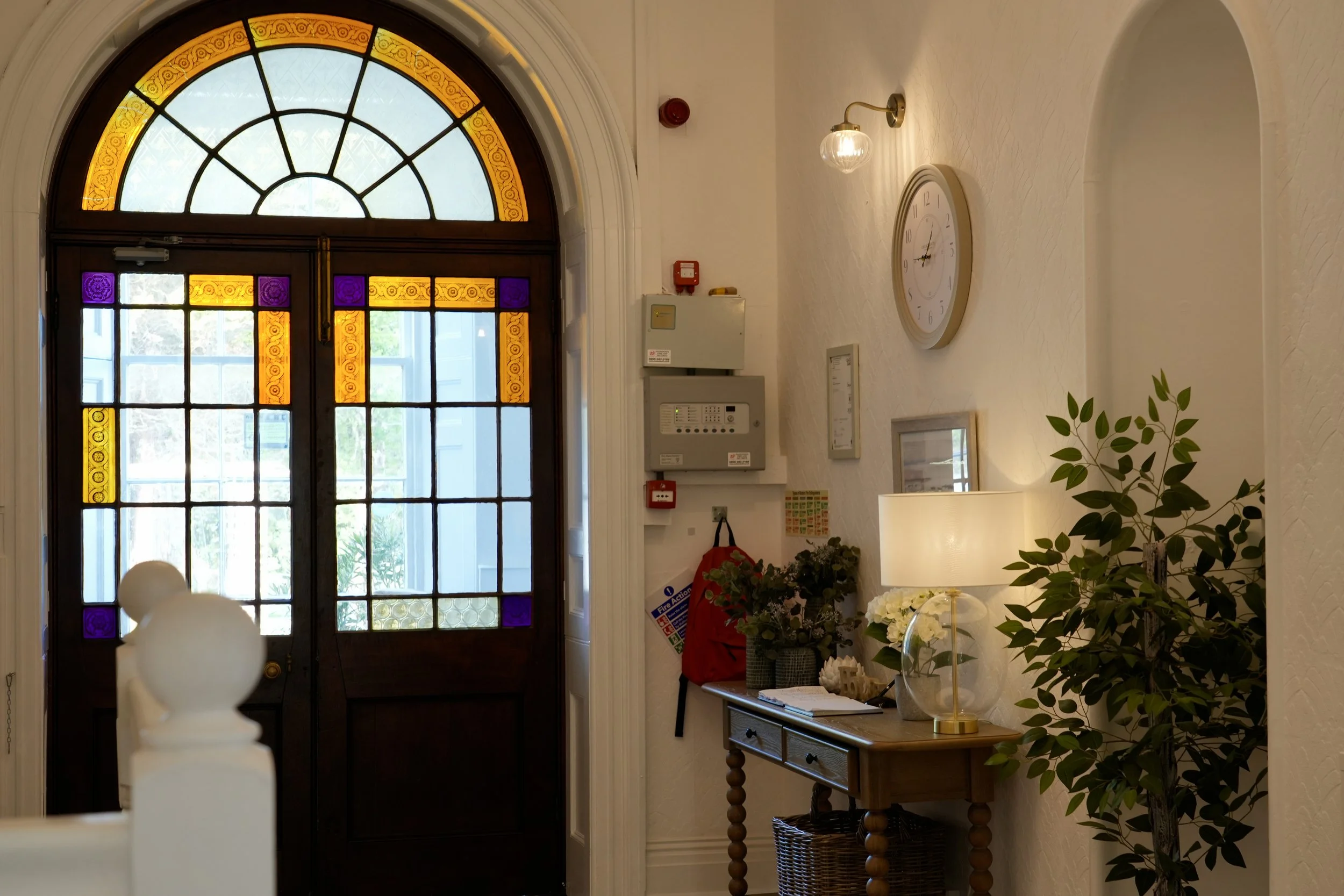 The image shows a hallway entryway with a wooden door featuring stained glass windows. There is a large round clock on the wall to the right of the door, and a brass wall sconce light above it. A small table is positioned underneath the clock, holdin