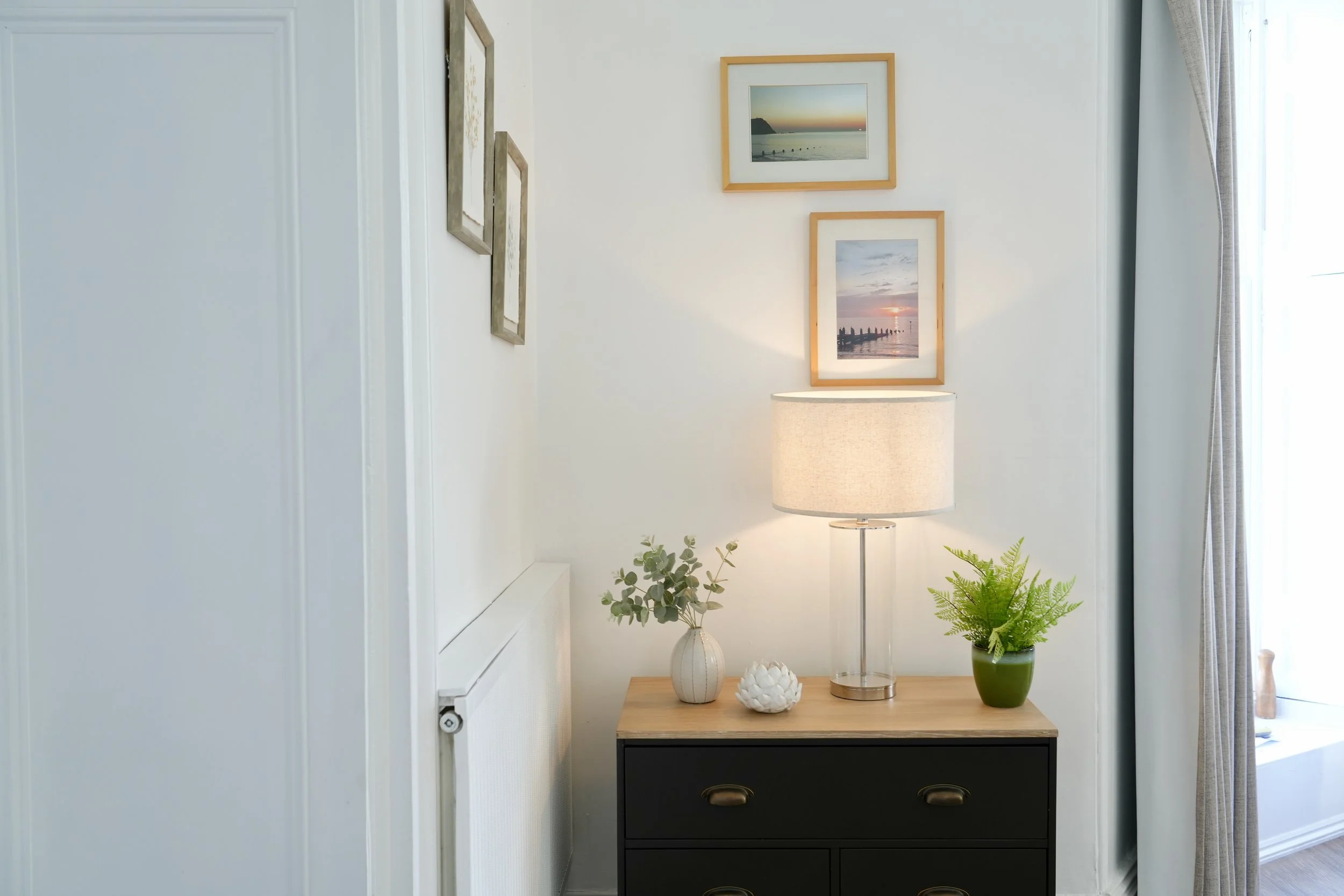 A side view of a black wooden dresser with three drawers, decorated with plants, ceramic decor, and a lamp on top, against a white wall adorned with framed pictures of coastal landscapes.