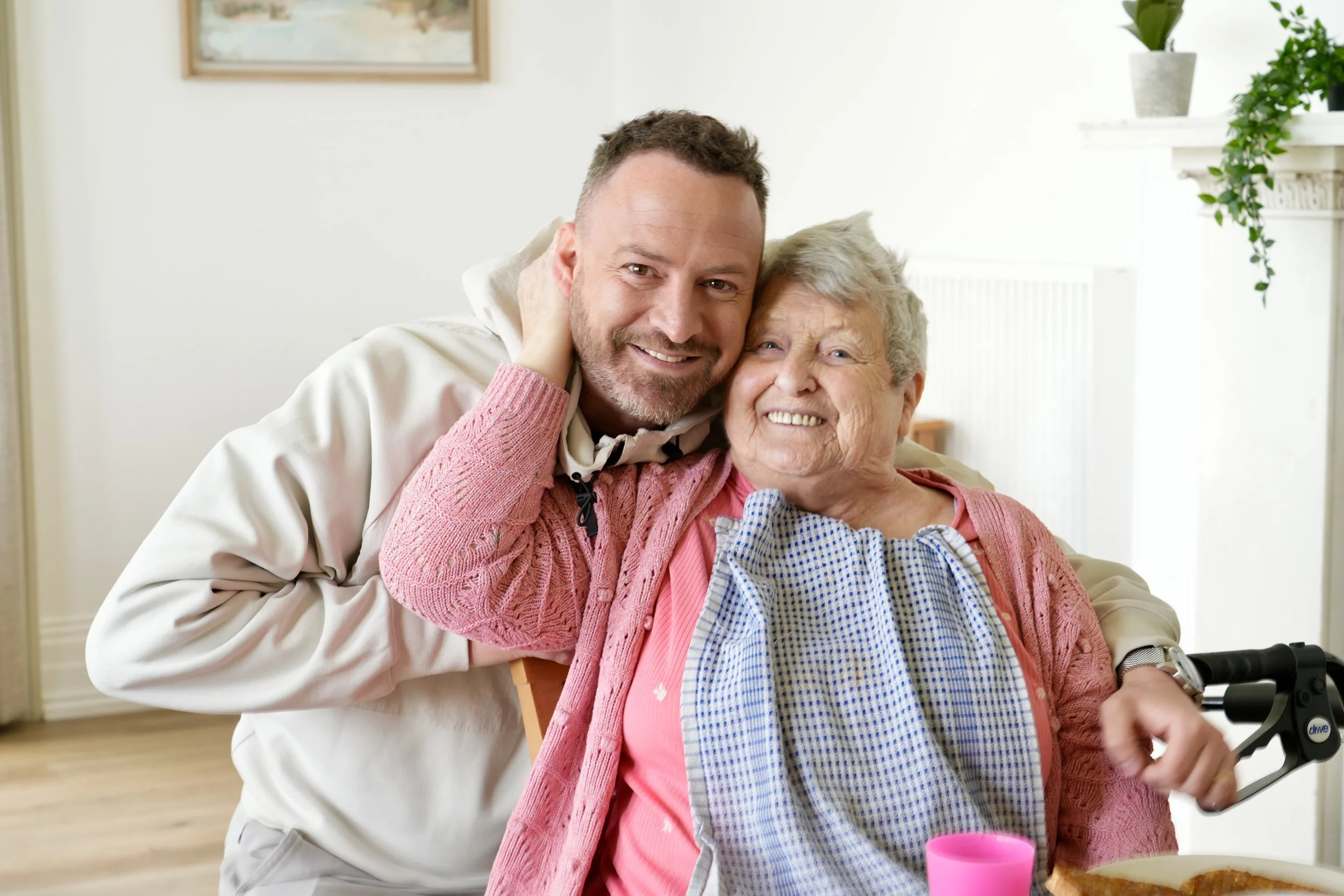 A man affectionately hugs an elderly woman who is in a wheelchair, both smiling happily in a room decorated with houseplants and a picture on the wall.