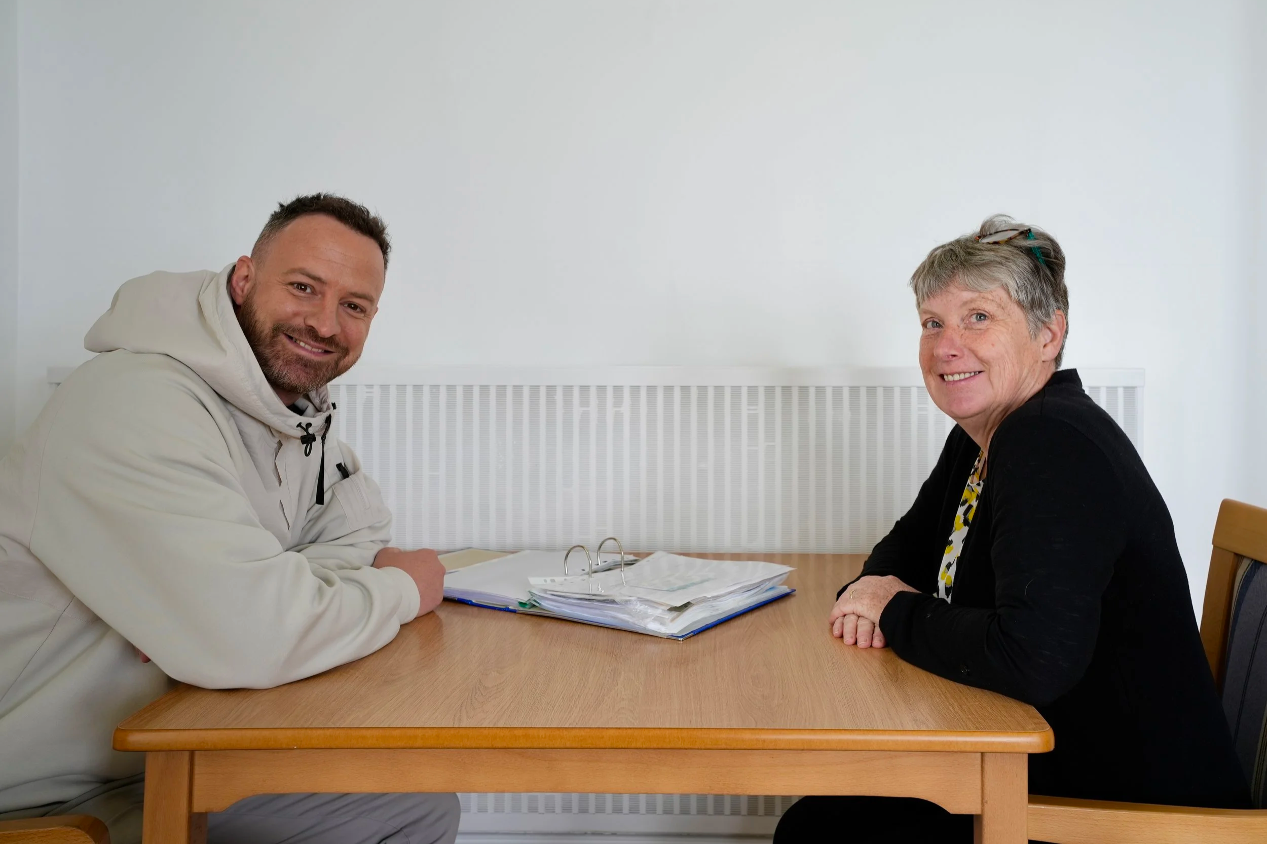 A man and woman sitting at a table with folders and papers, smiling at the camera, in a room with a white wall.