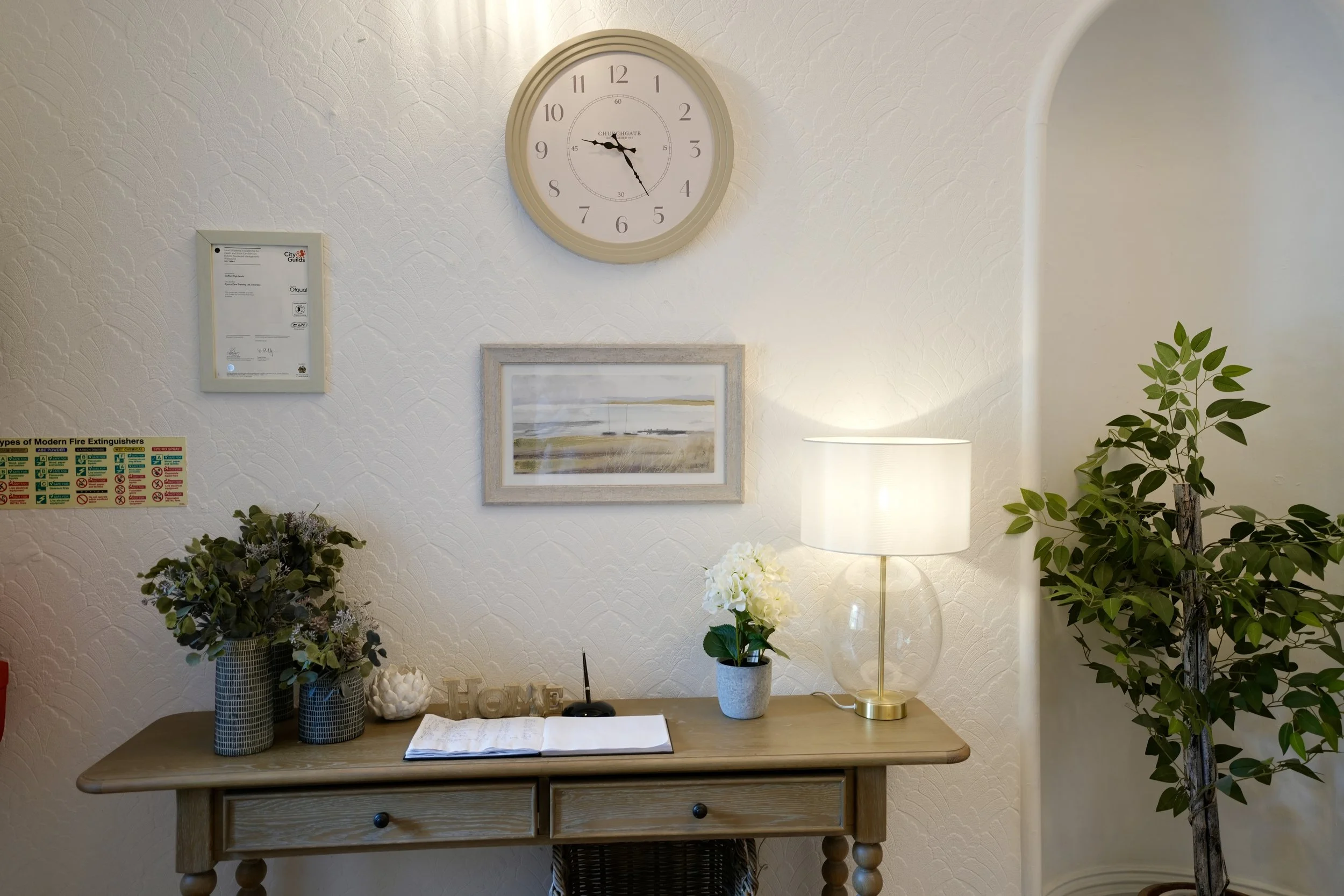 Decorative wall with a round clock, framed artwork, and various potted plants on a wooden table and nearby surfaces, with a lamp providing warm lighting.
