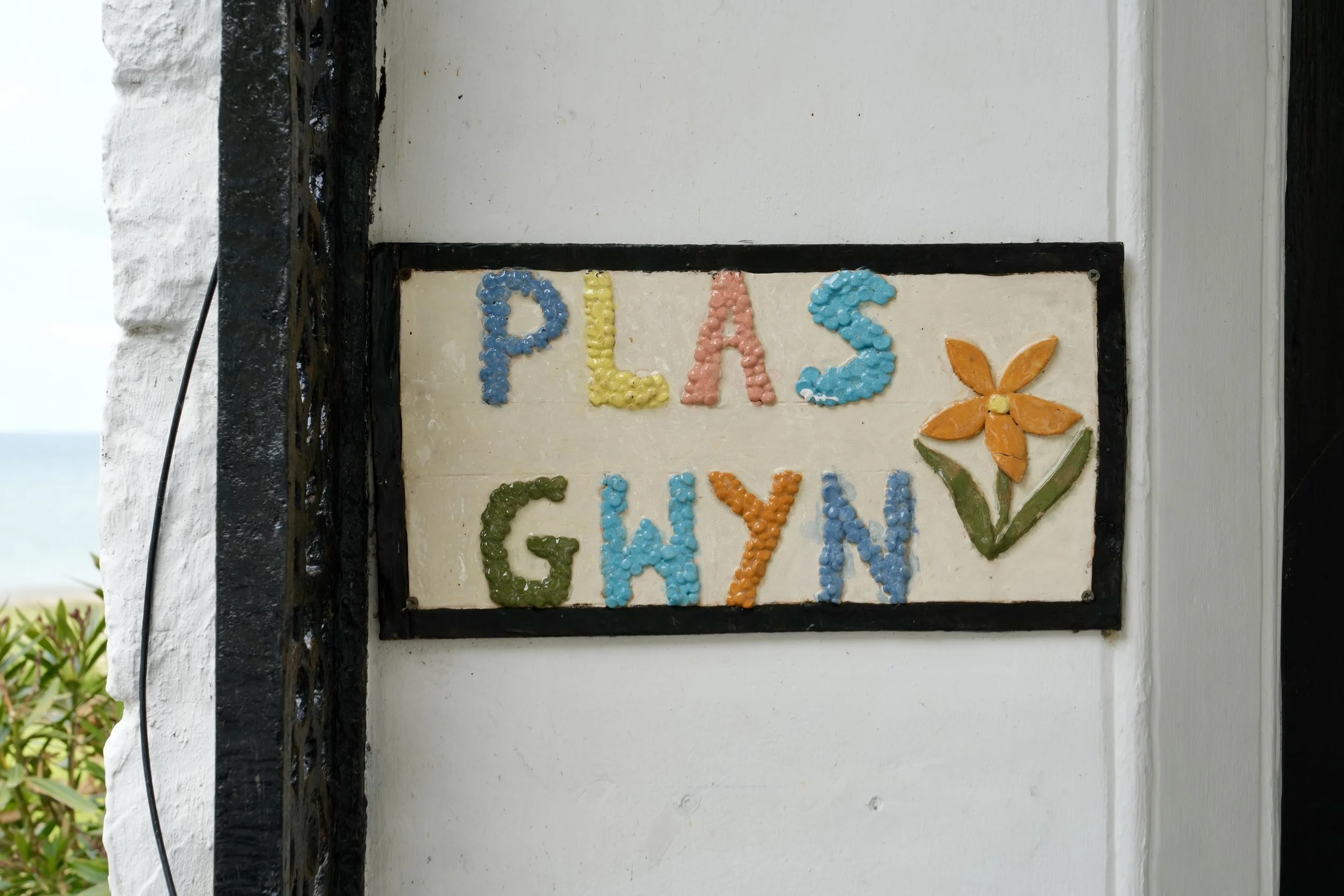Colorful sign with the words 'PLAS' and 'GIRL' made from painted stones, with a flower illustration on a white background, mounted on a wall.