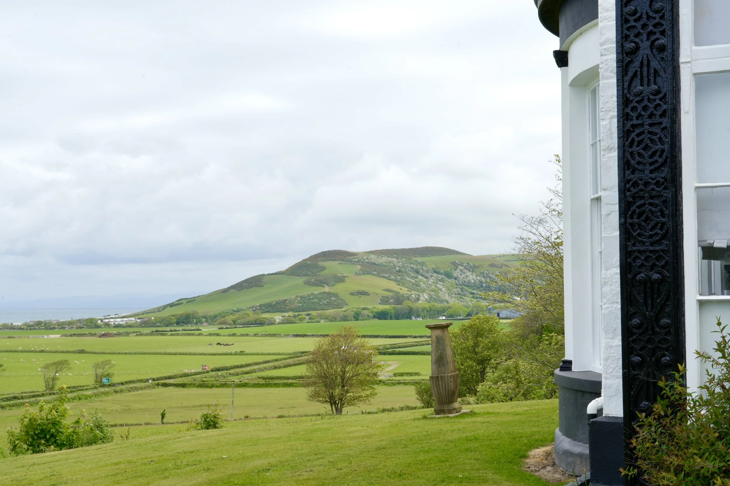 Scenic view of green rolling hills, fields, and trees with a cloudy sky in the background, partially seen from a house with white walls and decorative black ironwork.