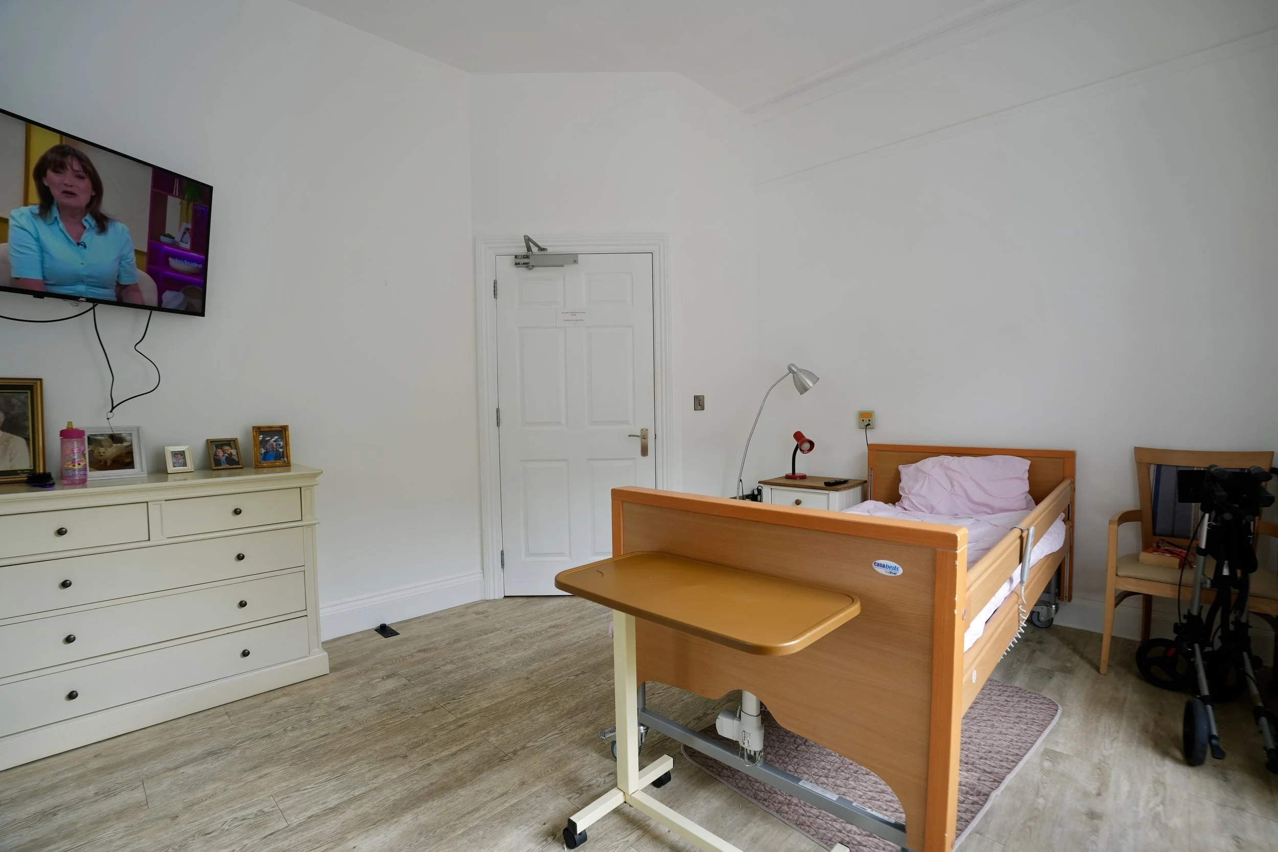 A hospital room with a wooden bed, pink sheets, and a bedside table with a lamp, a phone, and a red adjustable light. There is a TV on the wall showing a woman in a blue shirt, and several framed pictures on a white dresser. The door is white with a 