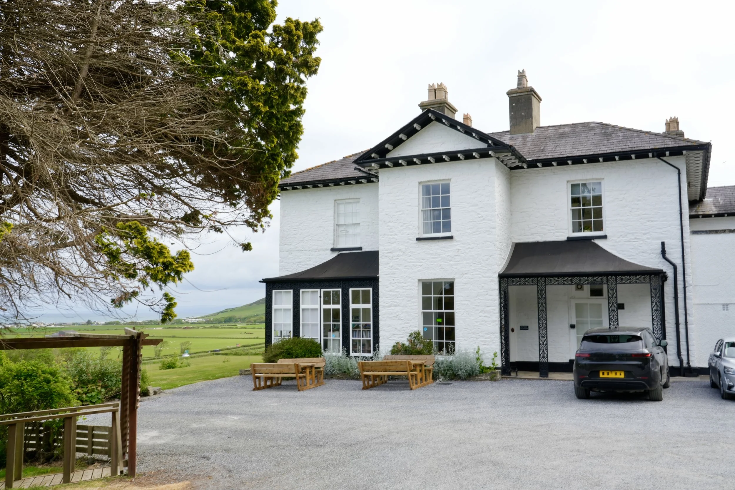 A white two-story house with multiple chimneys, set against a backdrop of green countryside and hills, with a gravel driveway, cars, outdoor seating, and surrounding greenery.