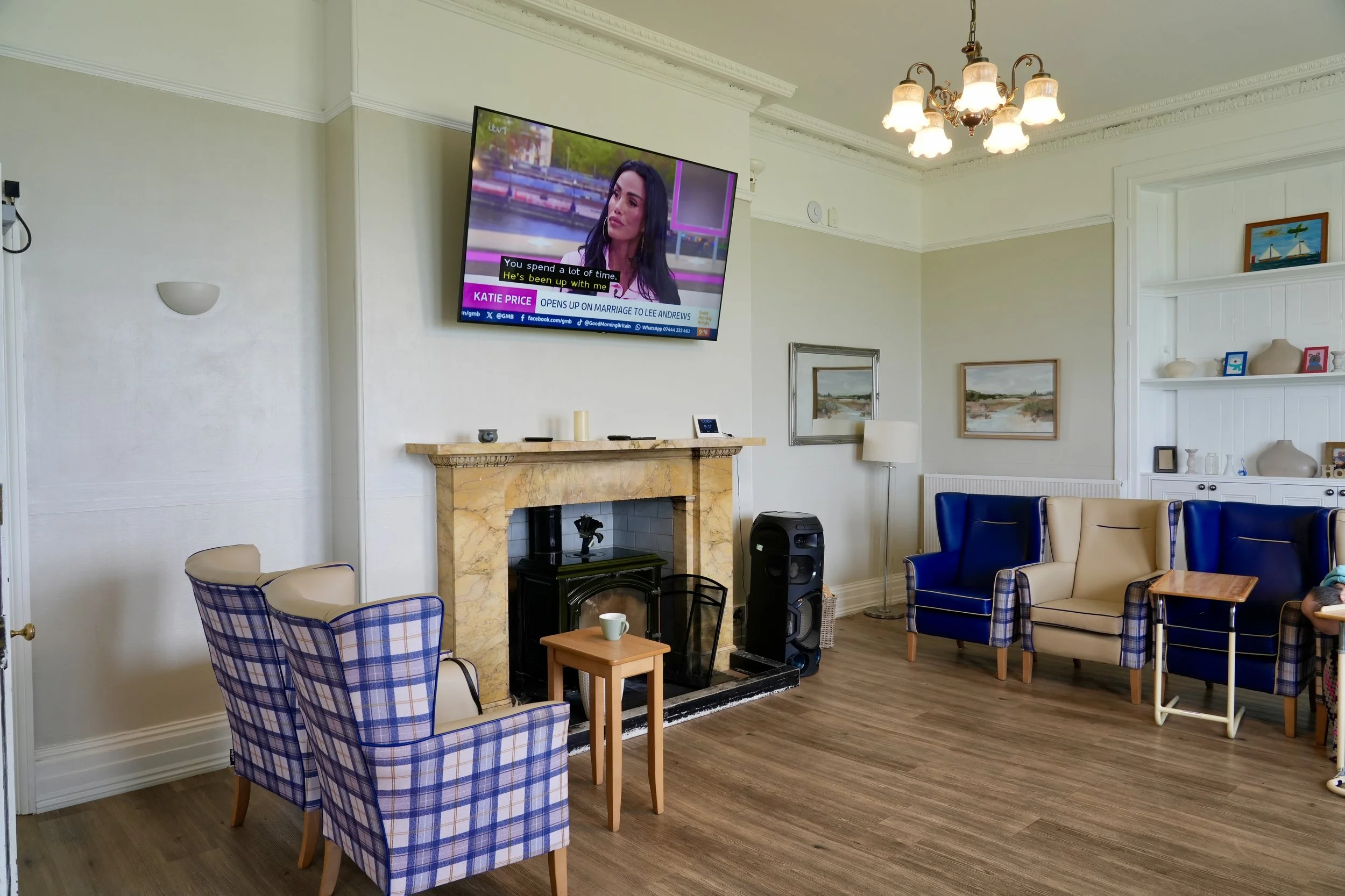 A cozy living room with a mounted flat-screen TV displaying a talk show, a marble fireplace, various chairs including plaid and beige armchairs, a small wooden side table with a coffee mug, a floor lamp, and decorative items on built-in white shelves