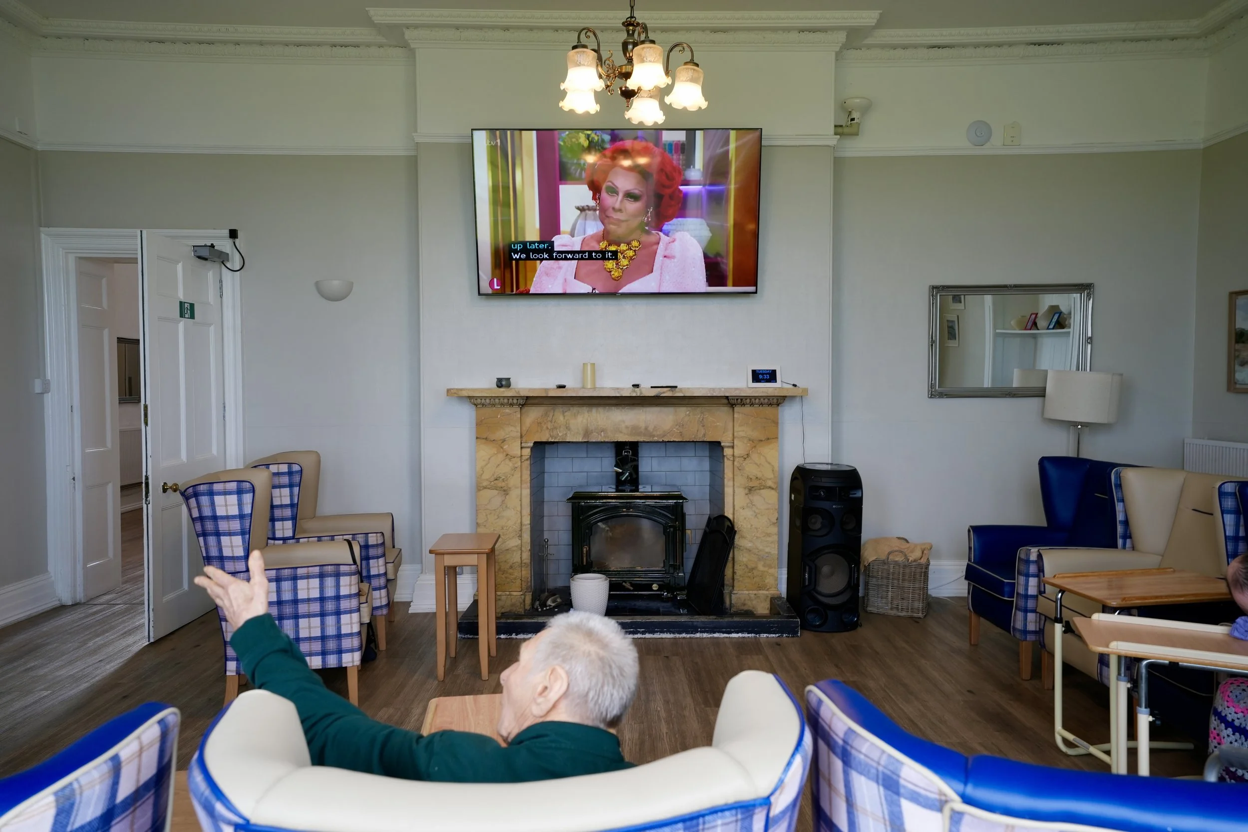 A living room with a fireplace, TV, and several chairs, where elderly people are seated watching a TV show featuring a woman with red hair and a pink outfit. A person's hand is visible in the bottom left corner, and the room has white walls and a cha